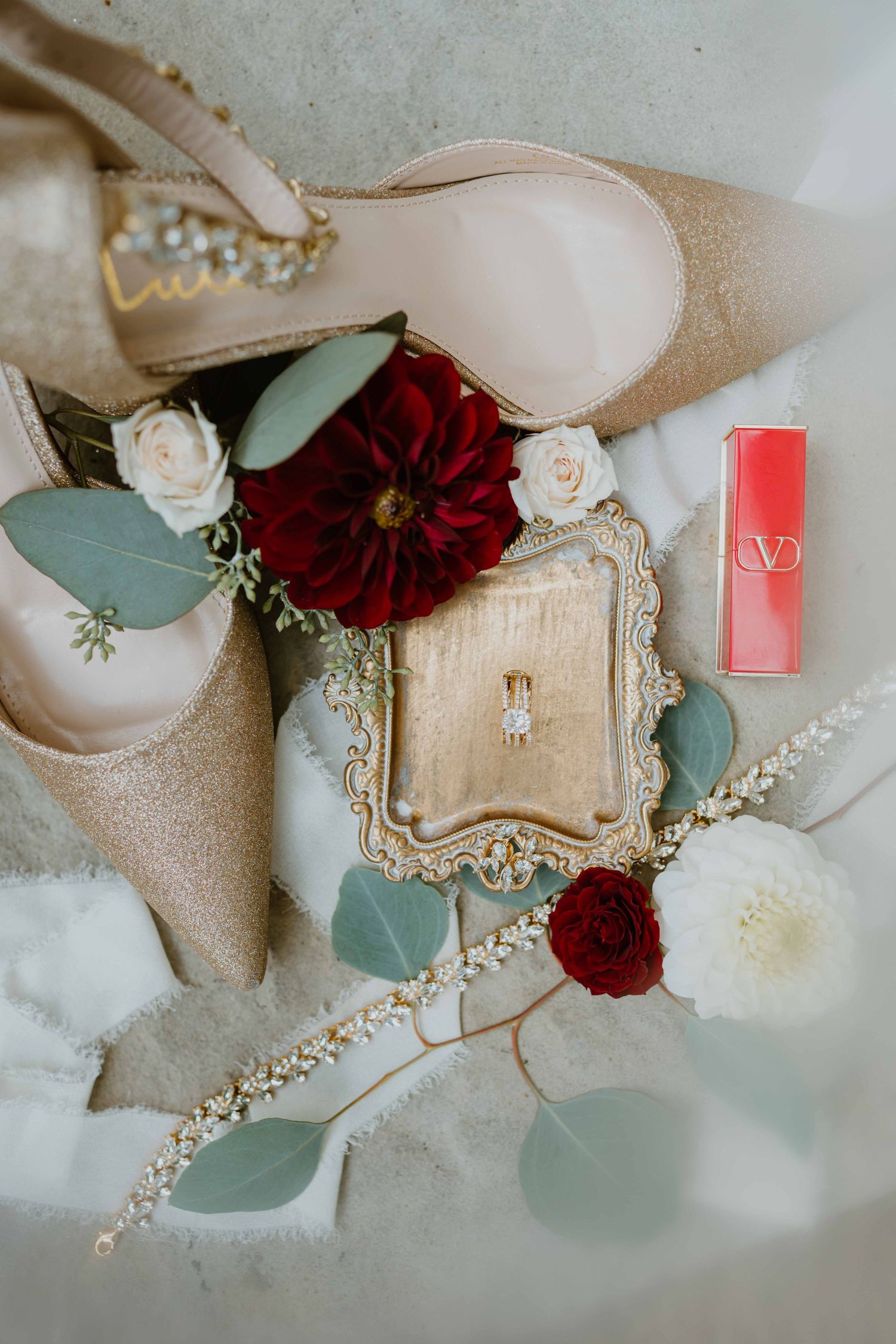 Bridal flat lay featuring gold glitter heels, diamond ring, red flowers, and Valentino lipstick.