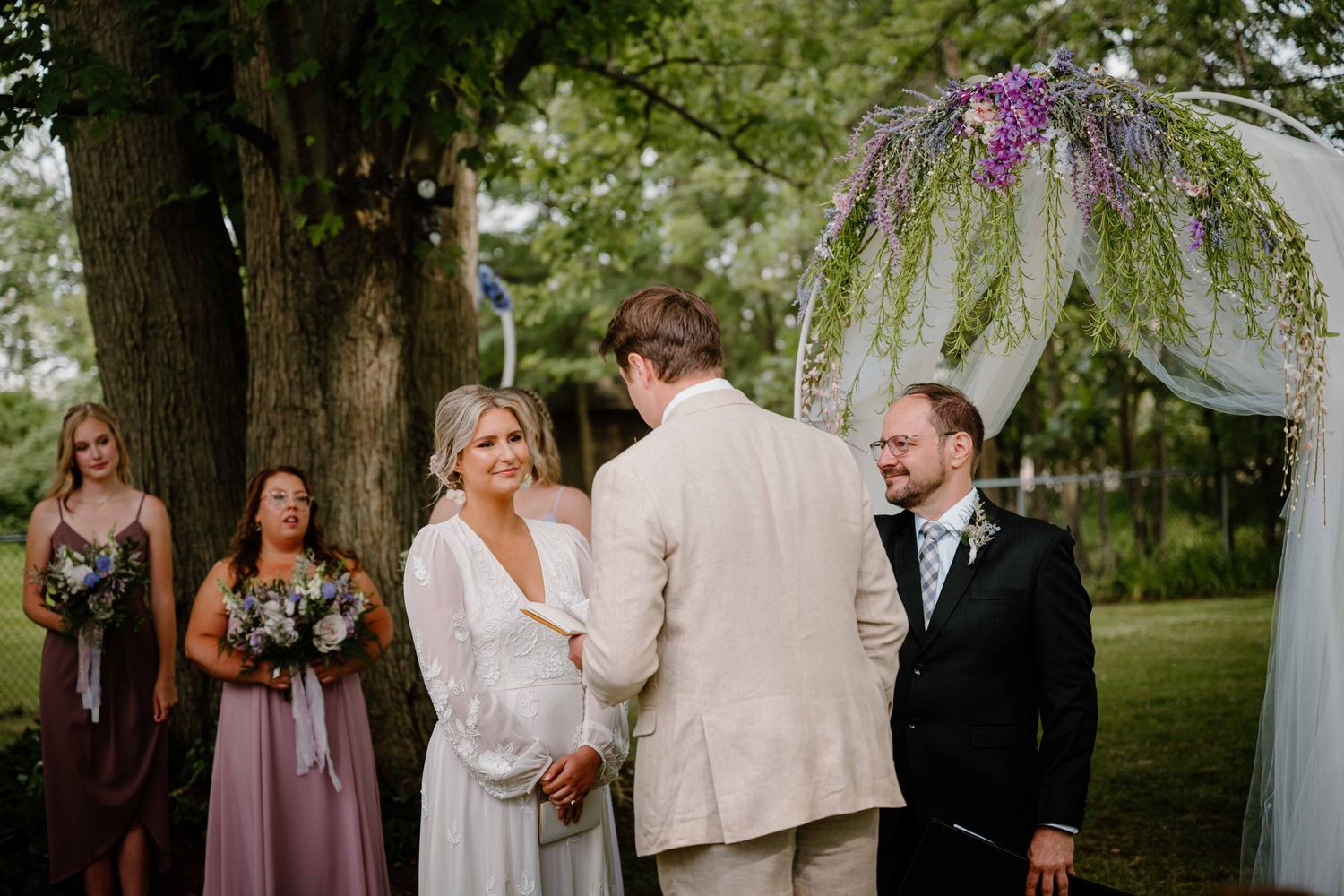 Bride laughs during outdoor wedding ceremony under purple floral arch surrounded by wedding party.