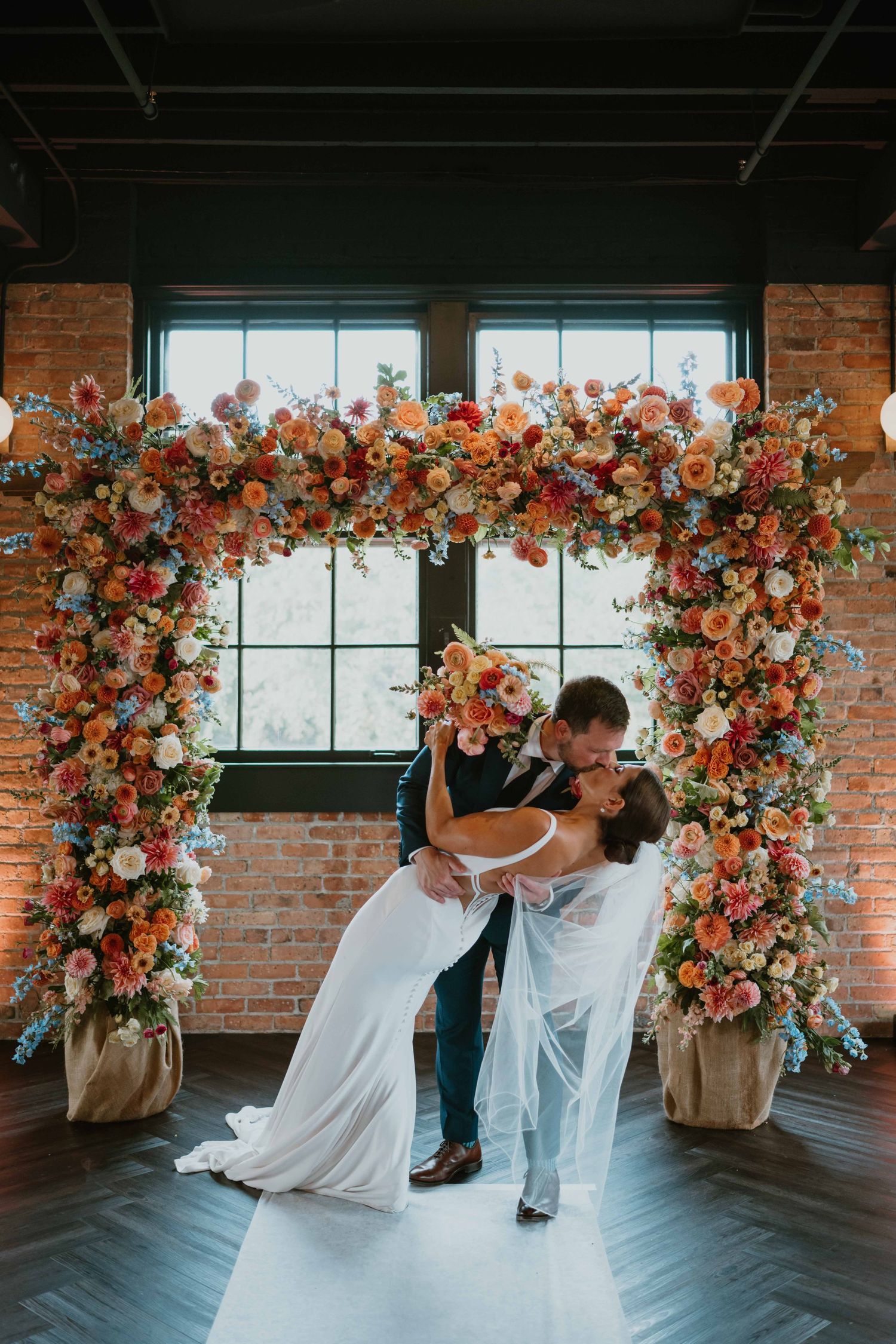 Bride and groom share a romantic dip kiss under a vibrant colorful floral arch at indoor wedding venue.