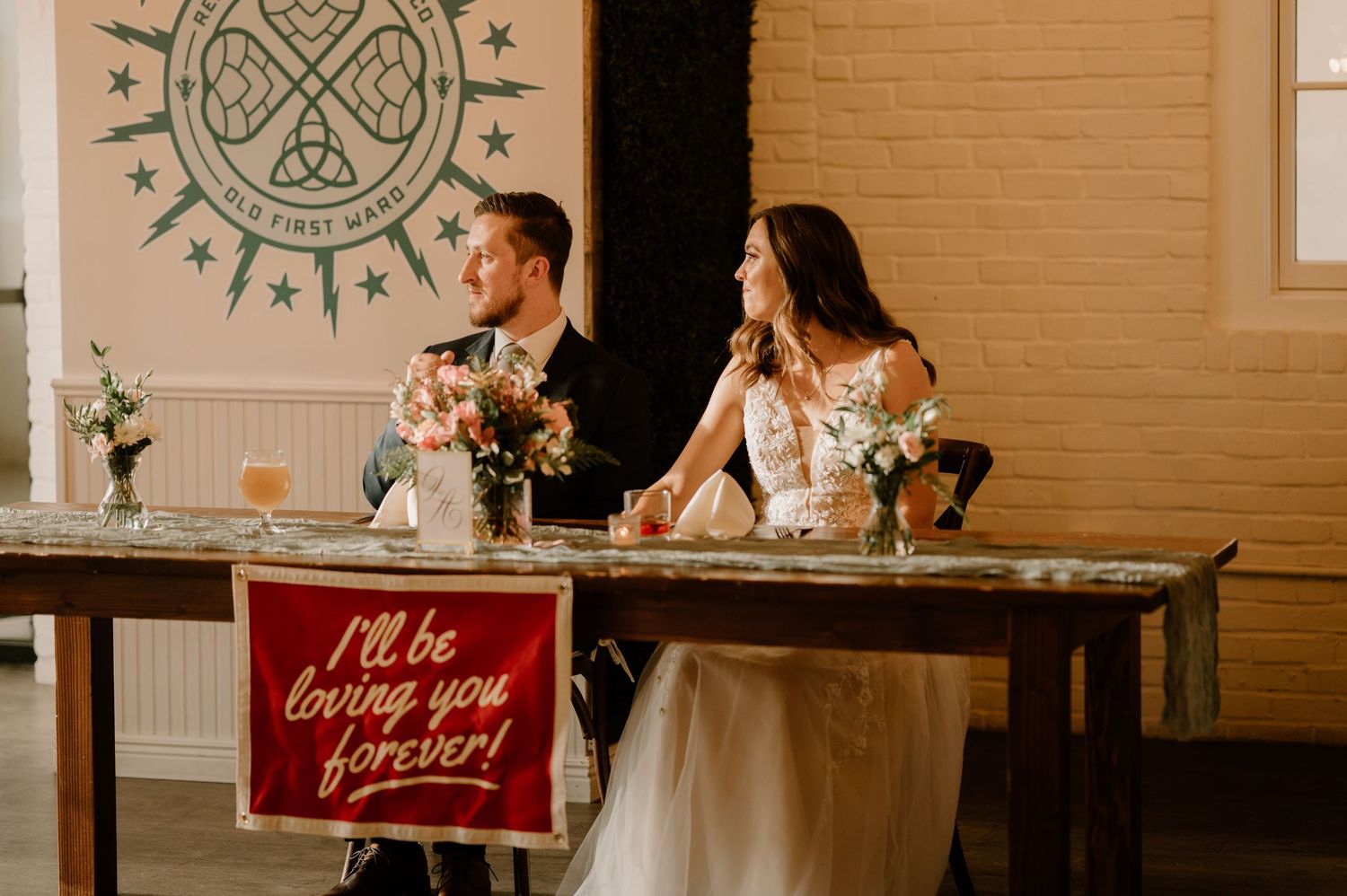 Bride and groom at sweetheart table with romantic banner reading I'll be loving you forever.