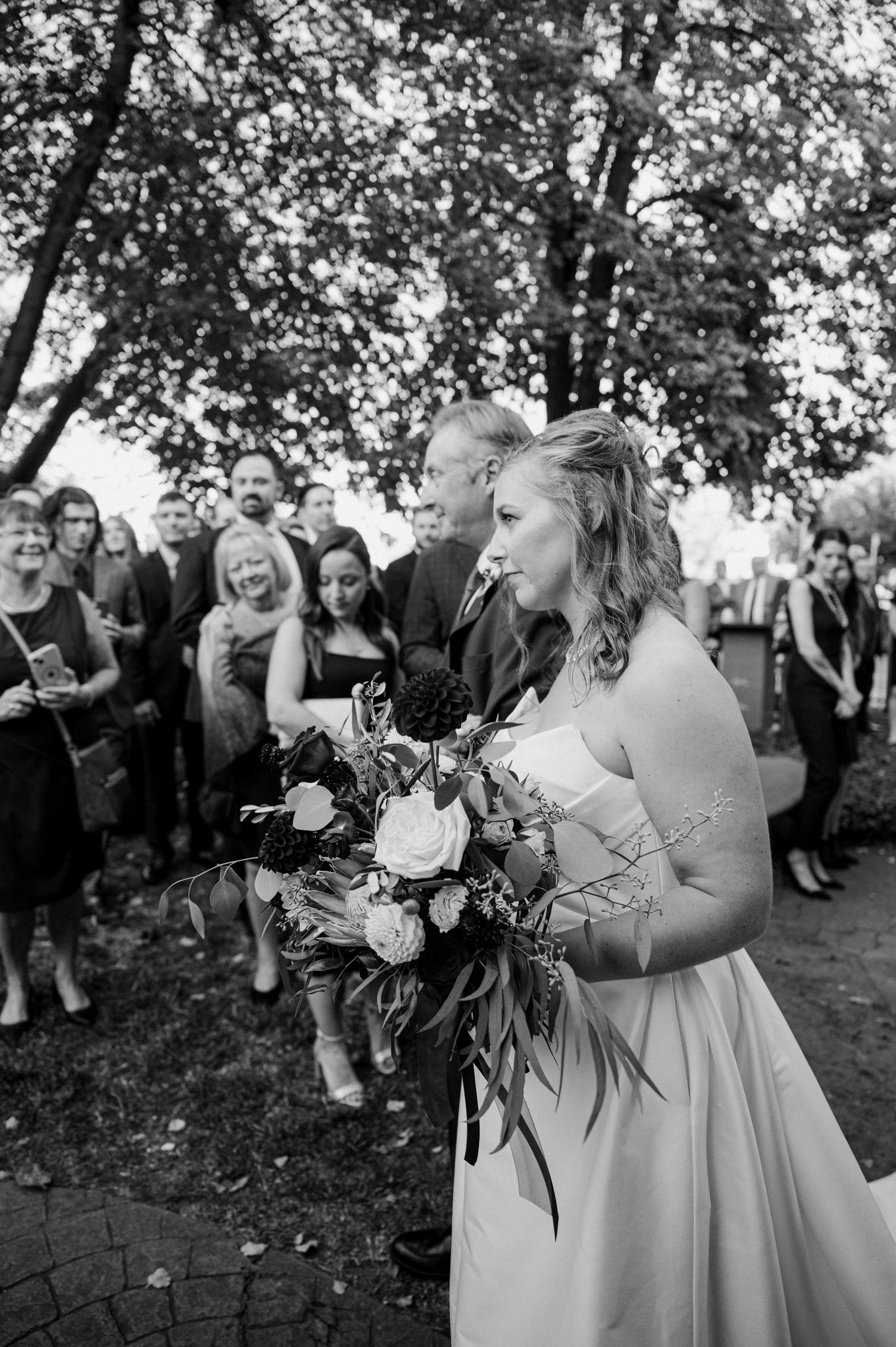 Bride holds a lush bouquet of white and dark flowers while guests gather behind her in black and white.