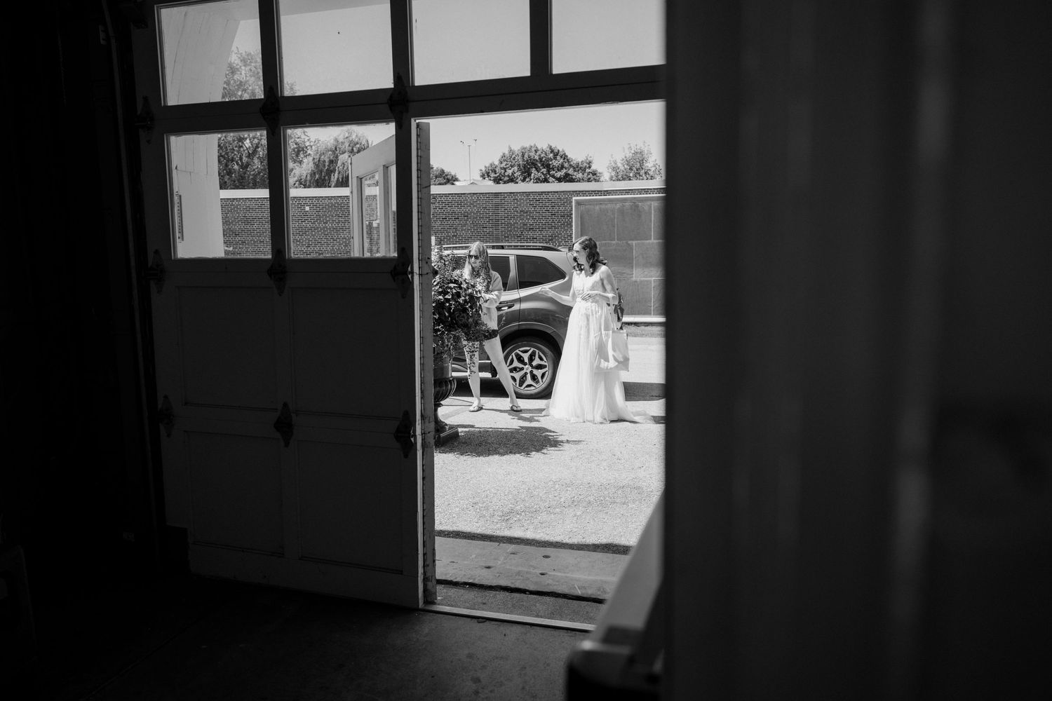 Bride in wedding dress viewed through open barn doors beside vintage car.