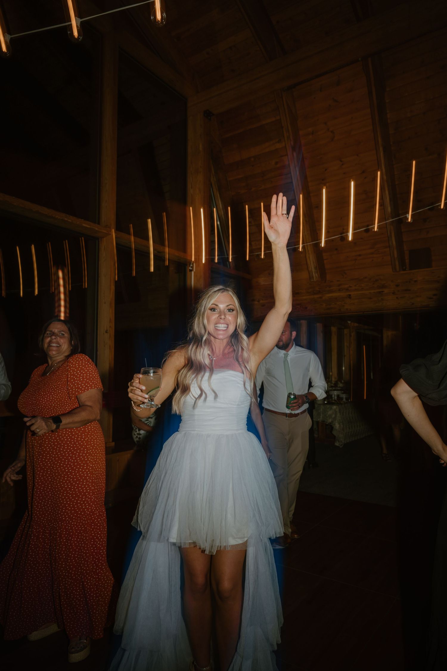 Bride in short white tulle dress raises hand while dancing at barn reception.