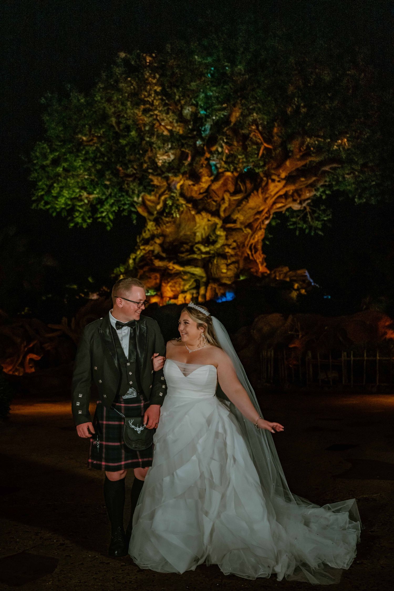 Bride and groom in Scottish kilt pose before the illuminated Tree of Life at Disney's Animal Kingdom at night.