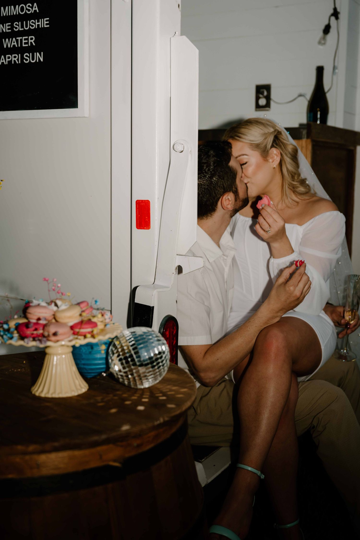 Bride feeds groom a treat near a donut display and disco ball at wedding reception.