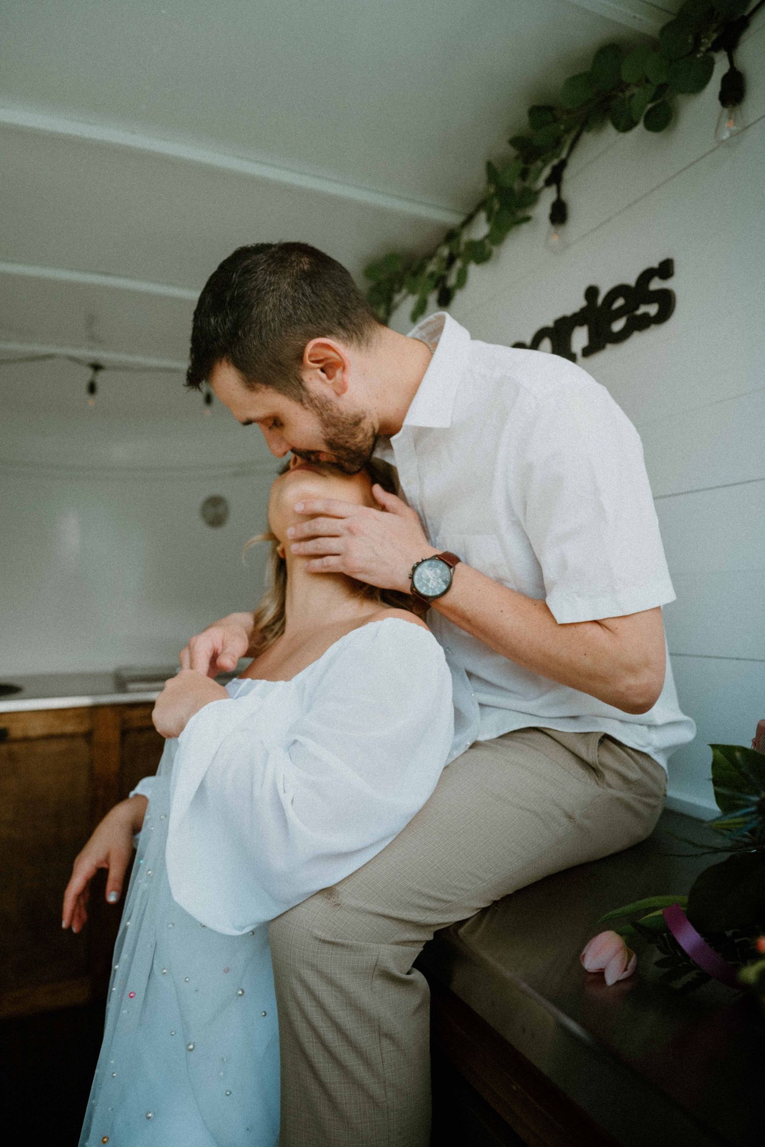 Groom in white shirt kisses bride on forehead while sitting inside a decorated bar truck interior.