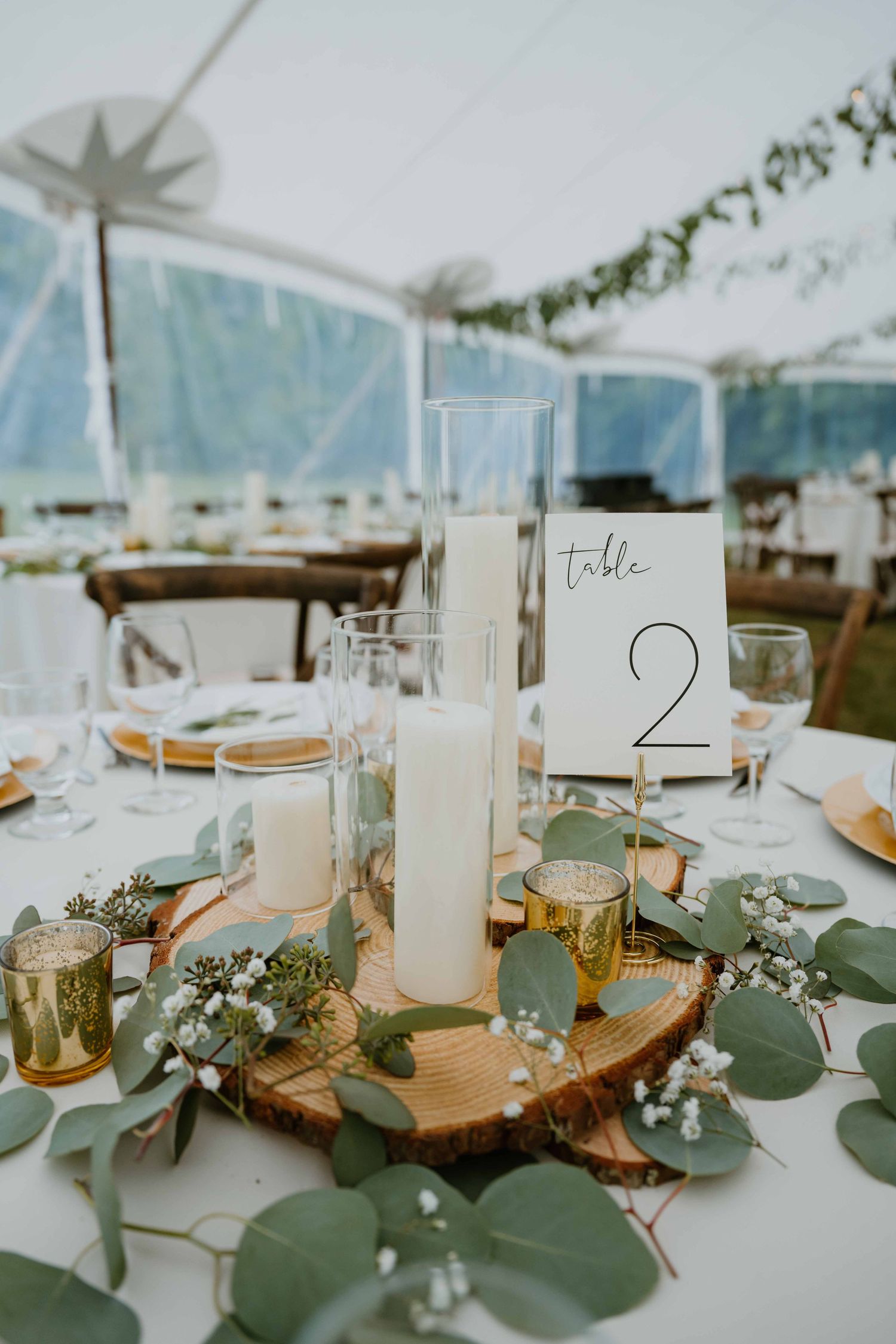 Elegant wedding table centerpiece with pillar candles, eucalyptus, and wood slice under tent.