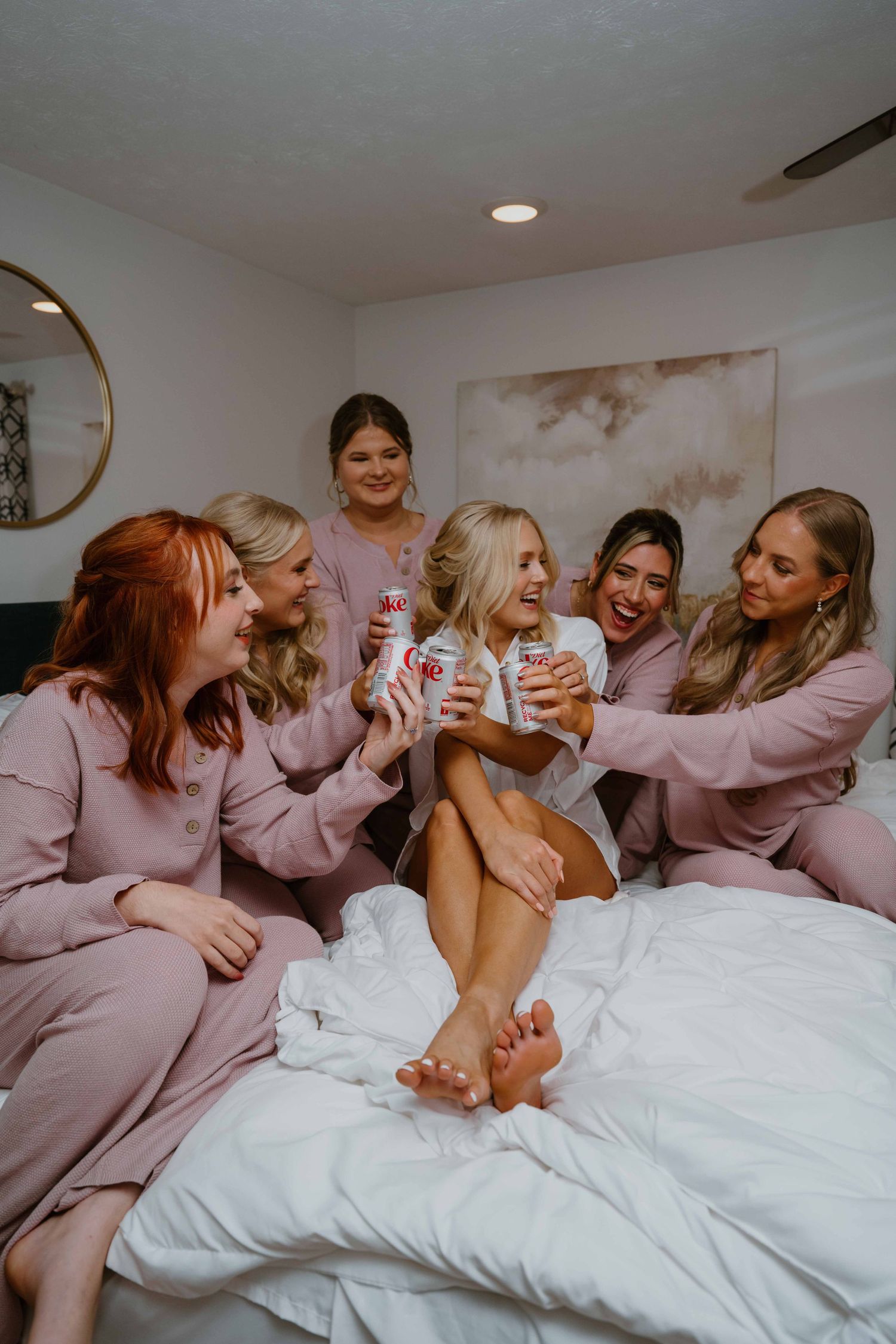 Bridesmaids in blush pink pajamas sitting on a white bed celebrating with canned drinks.