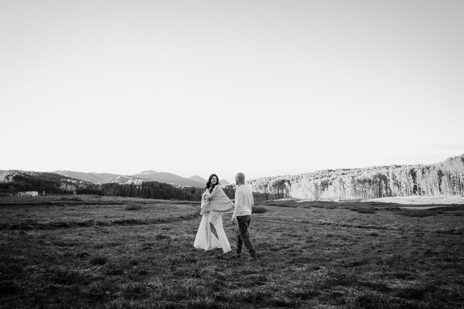 Black and white photo of bride and groom walking hand-in-hand across open mountain meadow.