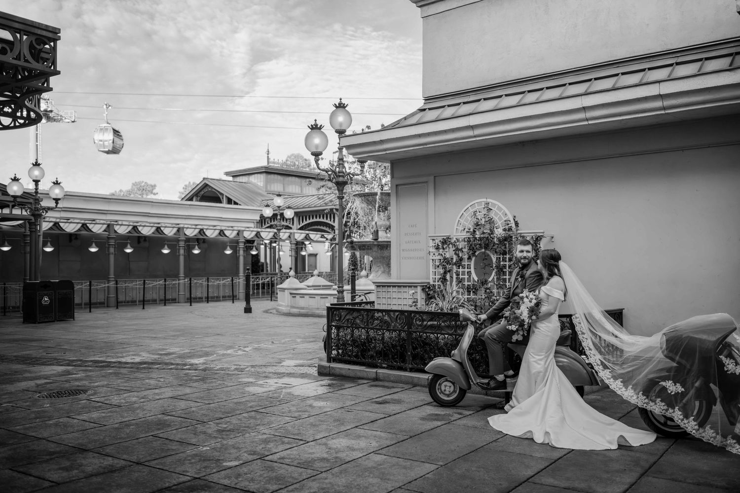 Black and white photo of bride and groom posing with vintage Vespa scooter in plaza.