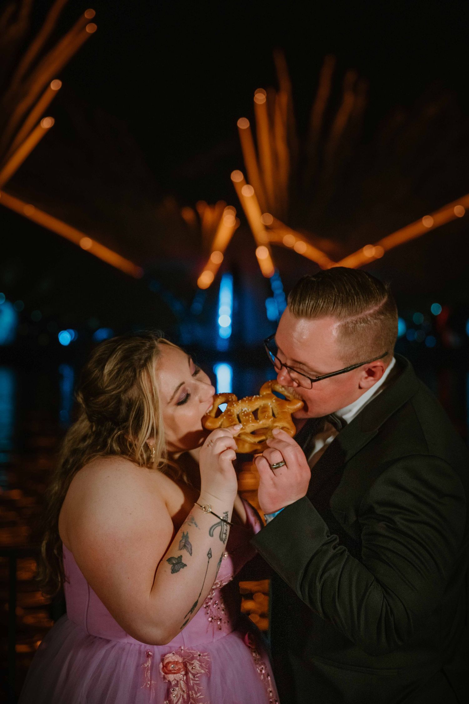 Bride and groom sharing a Mickey-shaped pretzel in front of colorful Disney World fireworks at night.