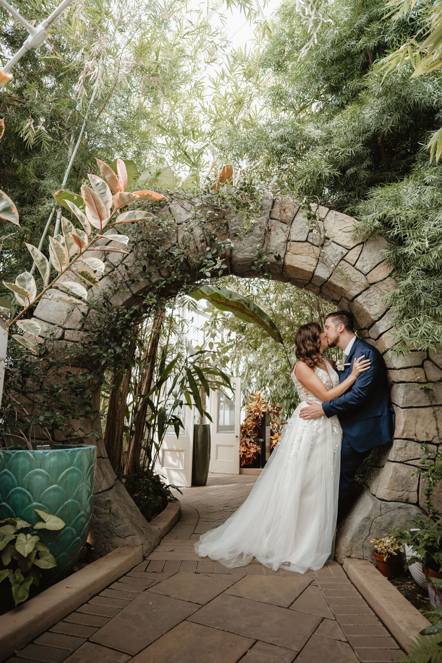 Bride and groom sharing a kiss under a stone arch surrounded by tropical greenery.