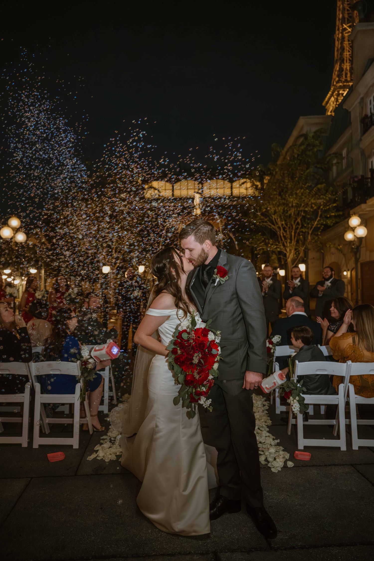 Wedding couple sharing first kiss as confetti rains down near illuminated Eiffel Tower replica.