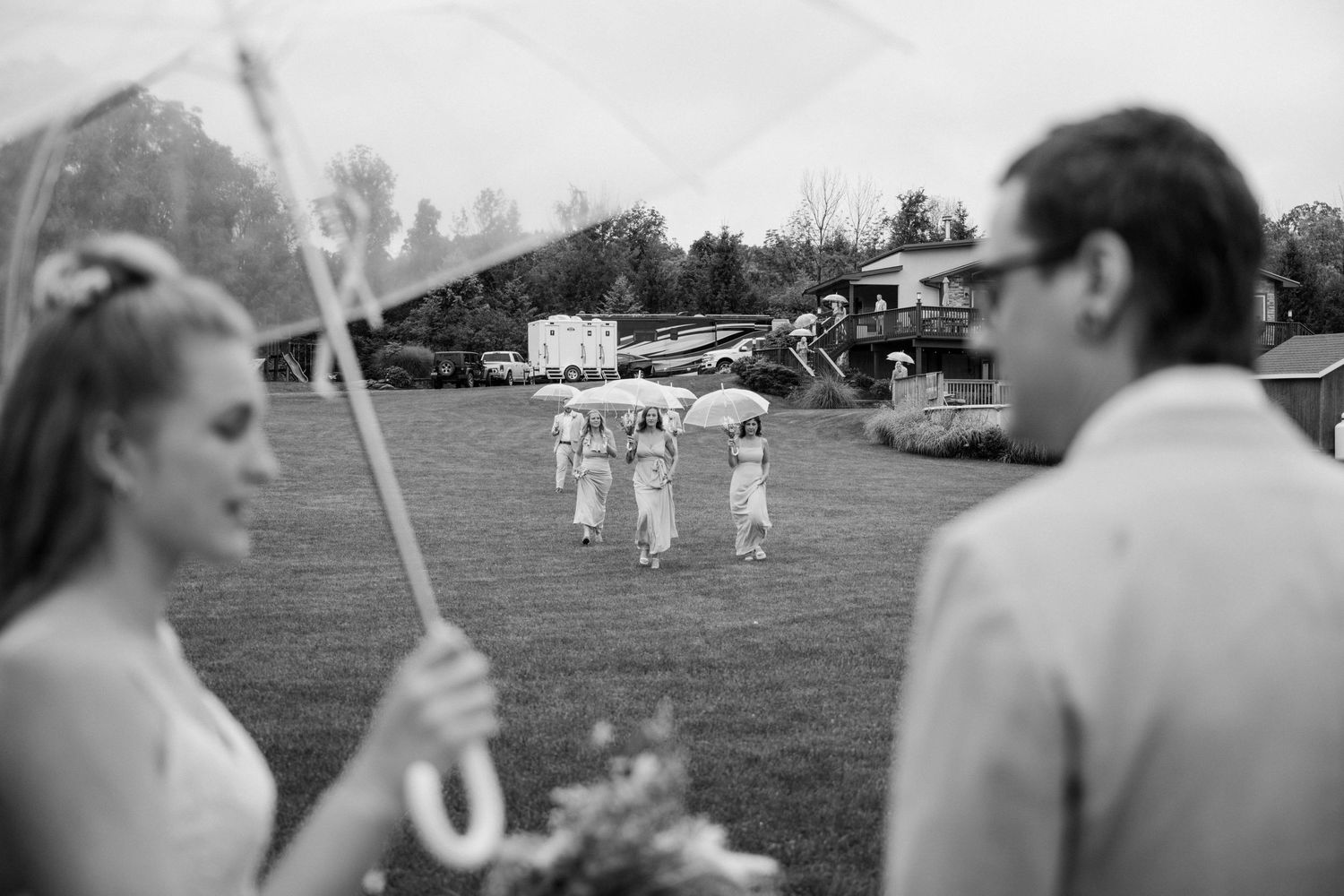Bride holds clear umbrella in rain while bridesmaids cross lawn with umbrellas at outdoor wedding.