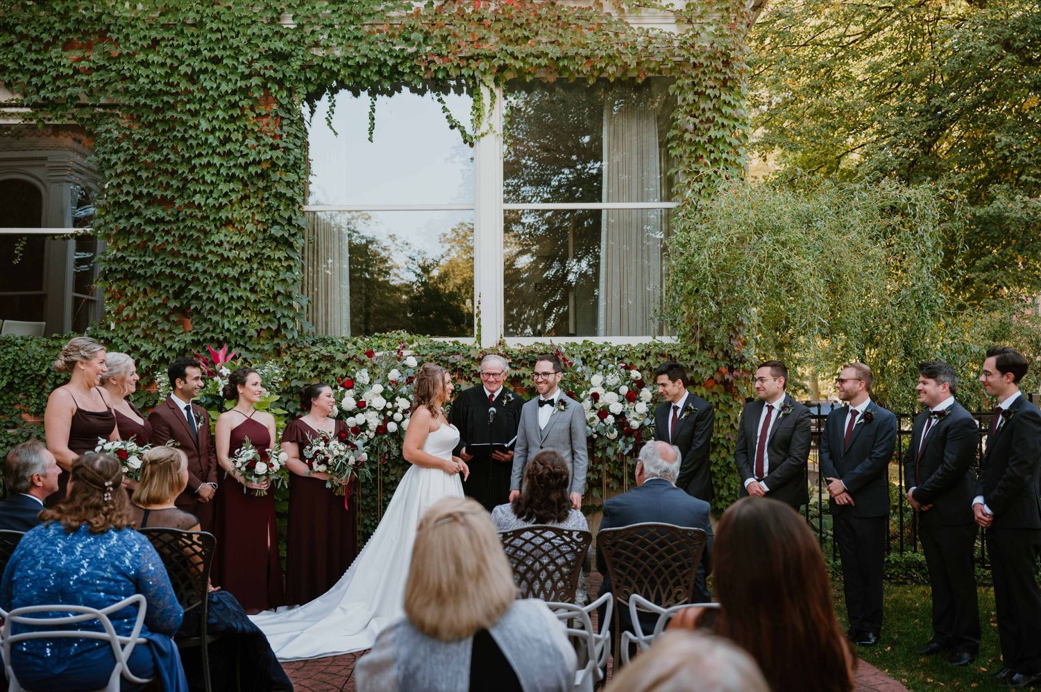 Wedding ceremony outdoors with ivy-covered wall backdrop, bridal party, and floral arch decorations.
