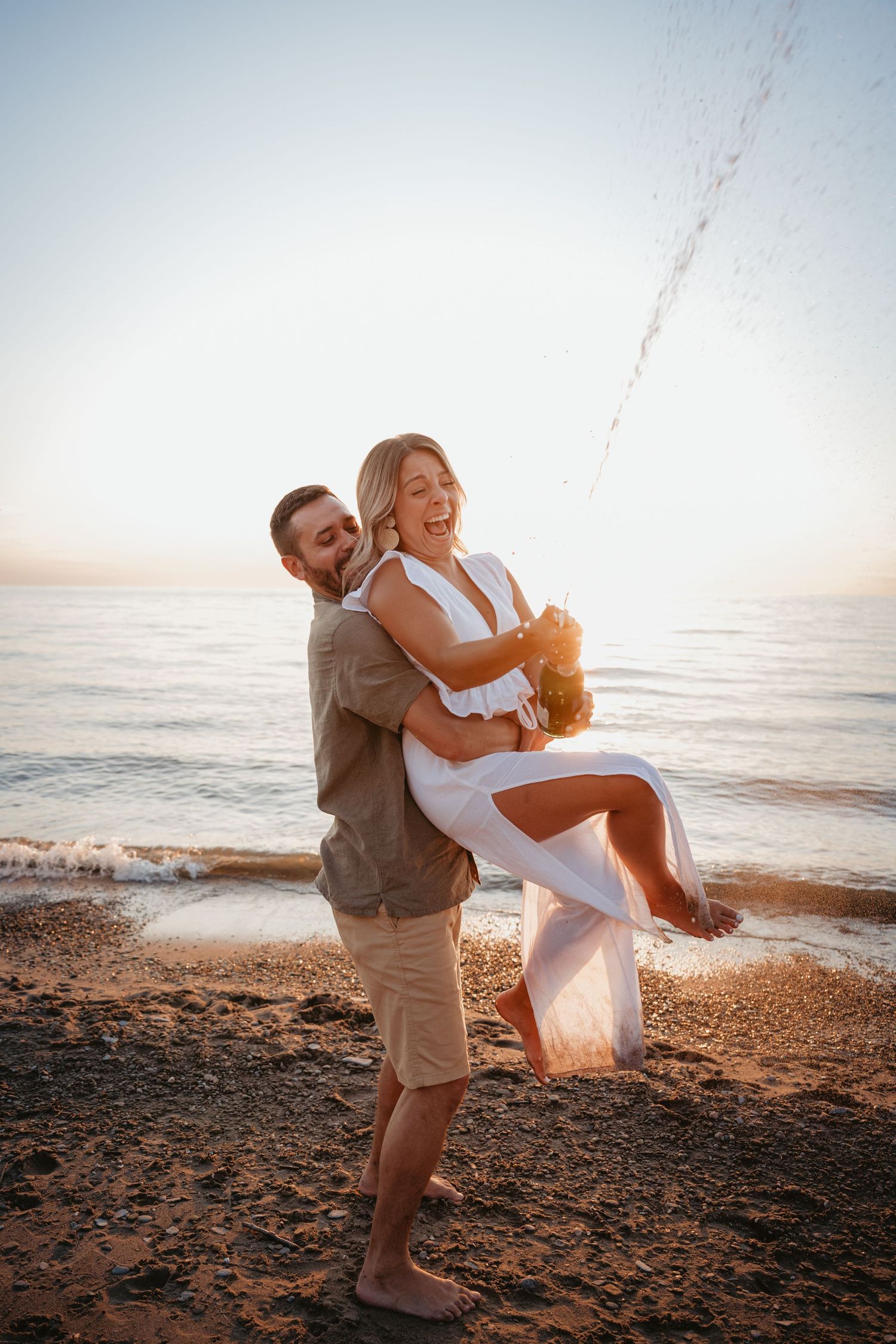 Man lifts laughing woman in white dress as champagne sprays during a joyful sunset beach celebration.