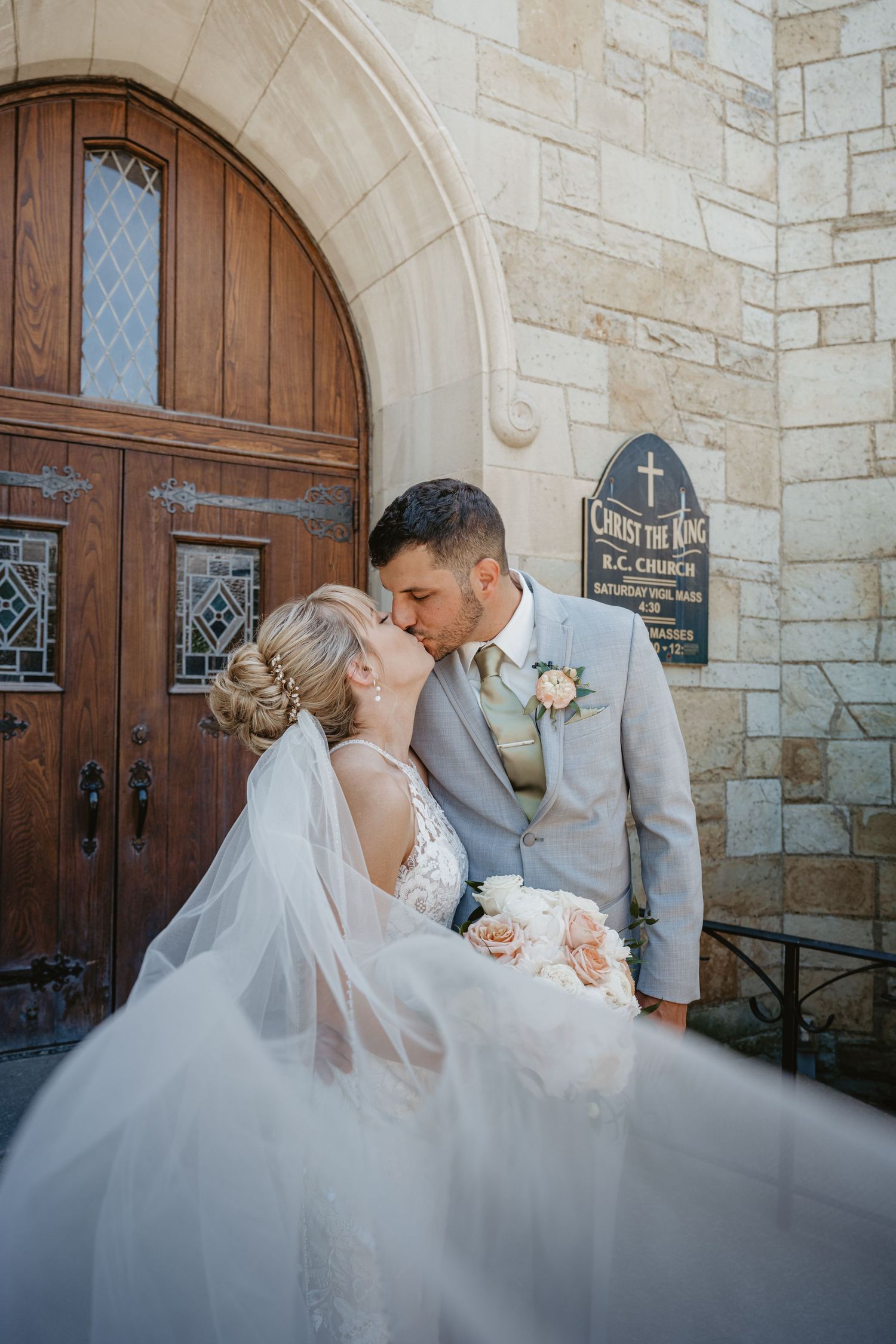 Bride and groom share a passionate kiss outside Christ the King RC Church, with veil flowing in the breeze.