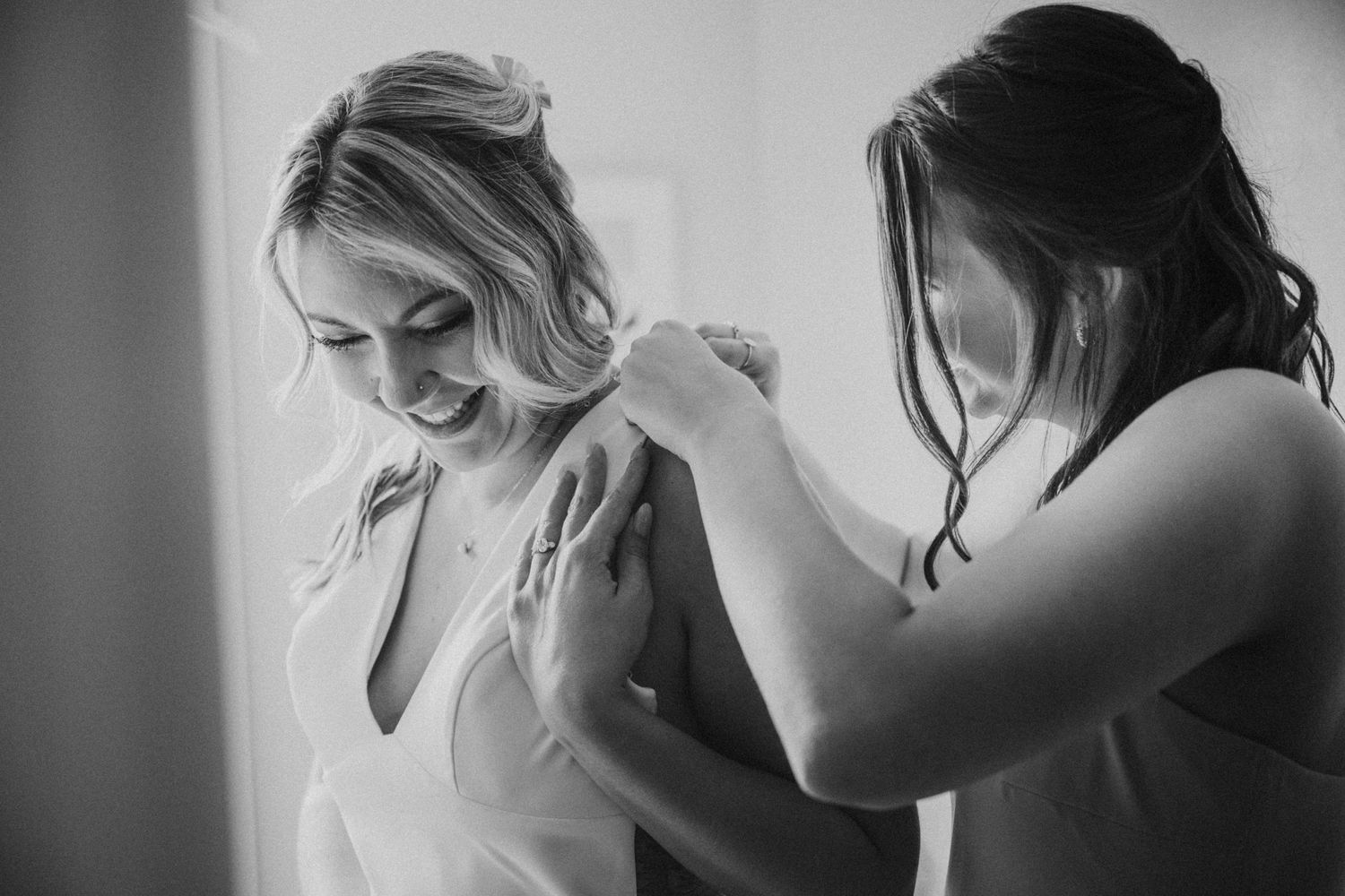 Bridesmaid helping smiling bride into her wedding dress in black and white photo.