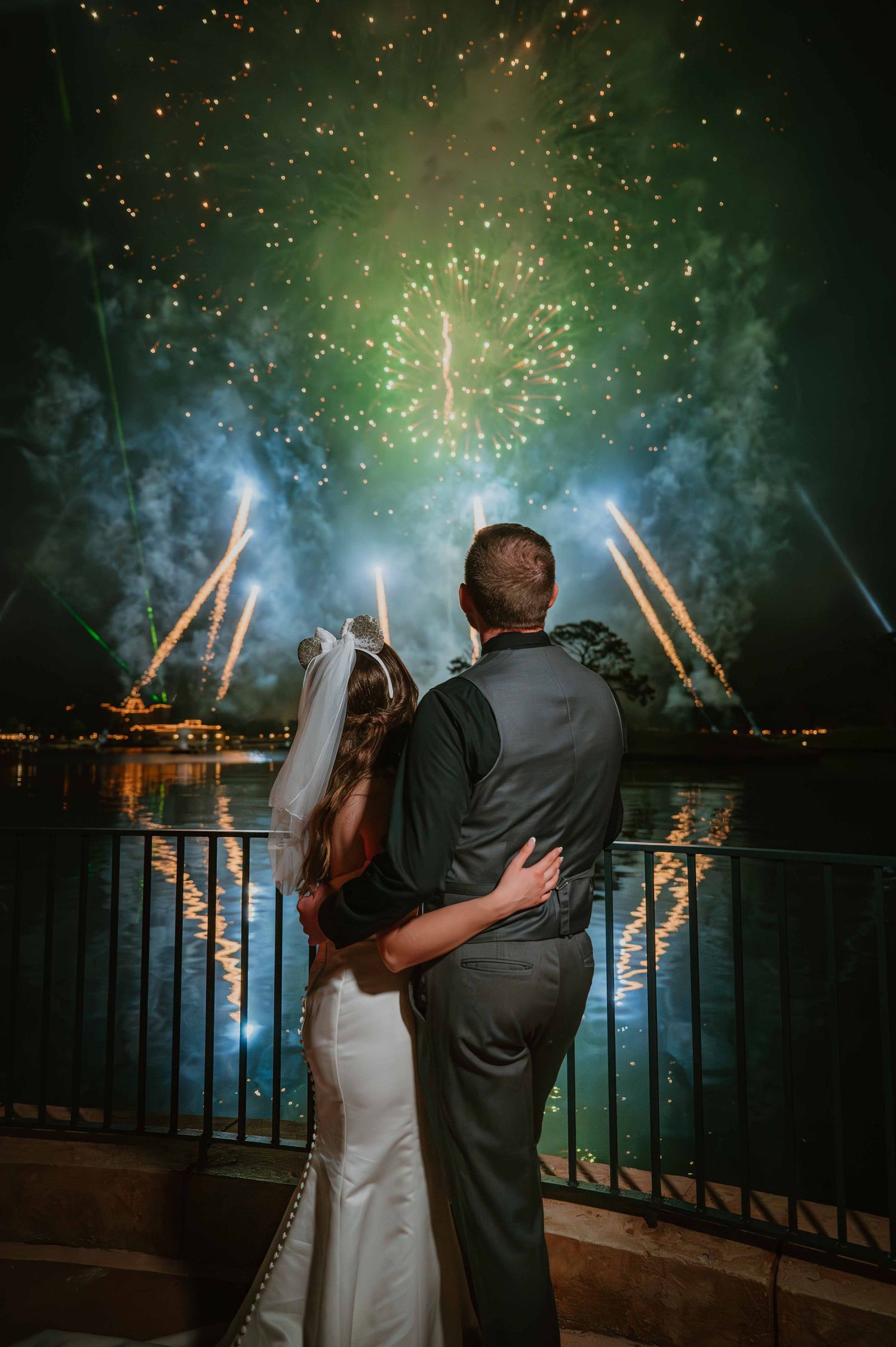 Bride and groom embracing while watching stunning green and gold fireworks over lake at night.