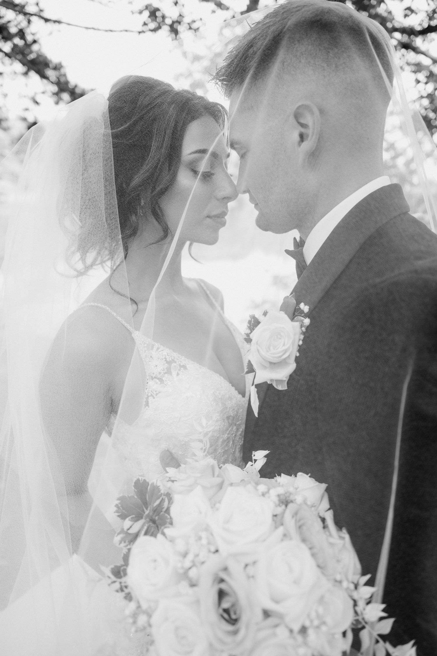 Bride and groom touching foreheads under veil with wedding bouquet in black and white.