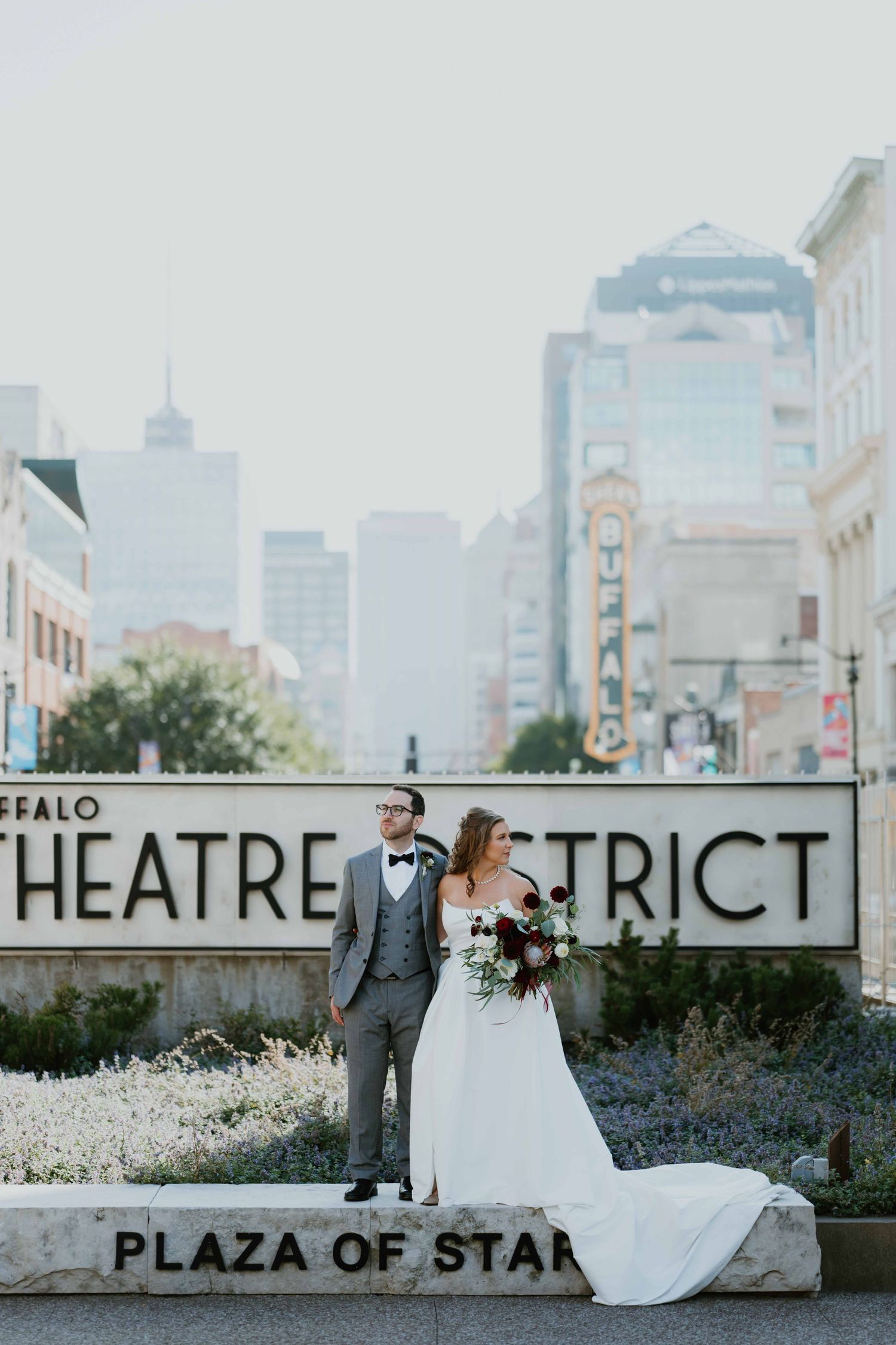 Bride and groom pose at Buffalo Theatre District Plaza of Stars sign with city skyline backdrop.