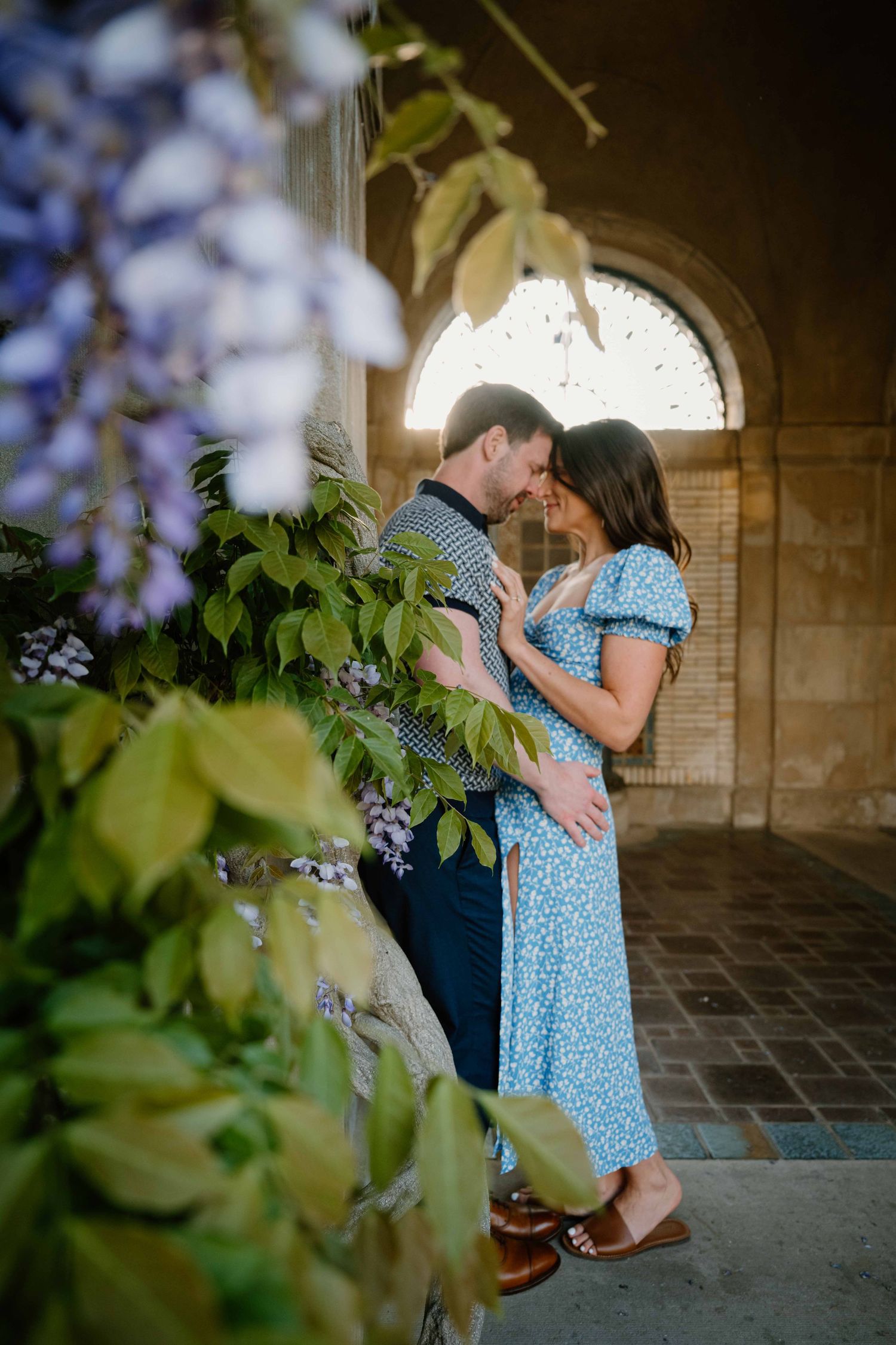 Romantic couple embraces among purple wisteria blooms framed by a stone archway in soft natural light.