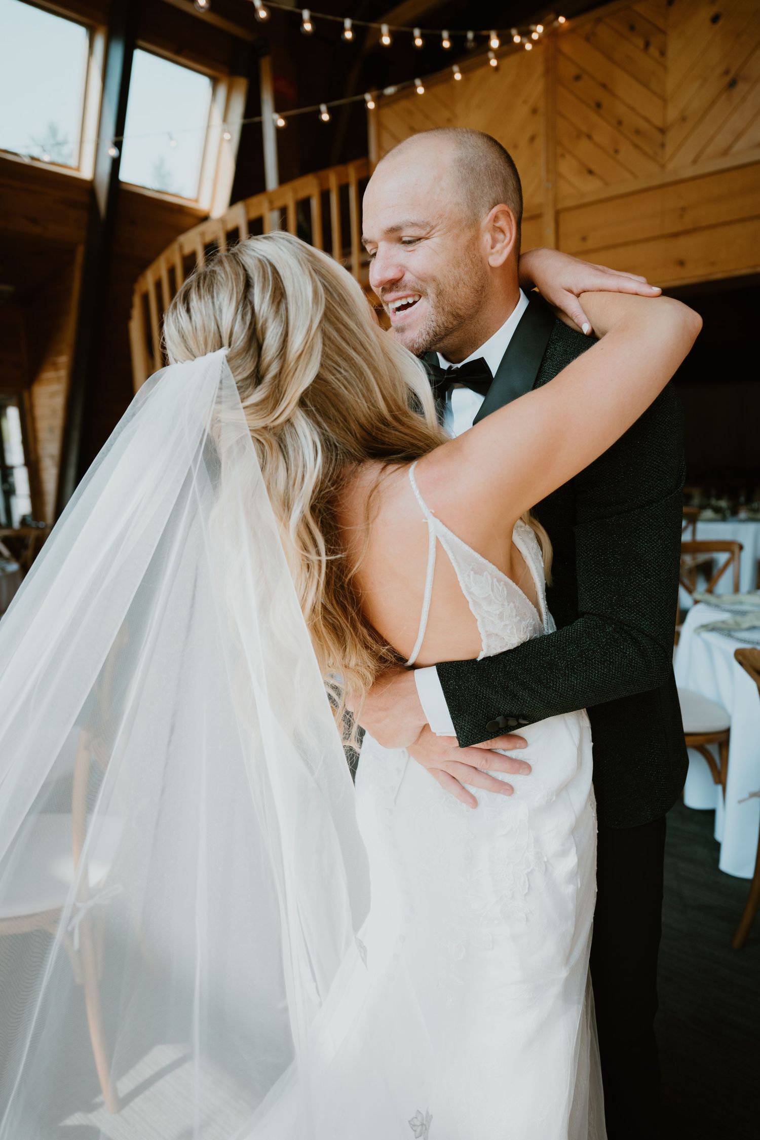 Groom smiling joyfully as bride embraces him during first look in a rustic barn venue.