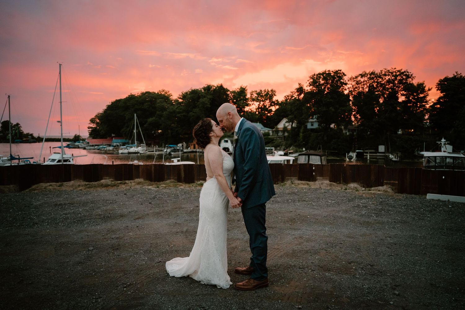 Bride and groom share a kiss against a breathtaking pink and orange sunset sky at waterfront marina.