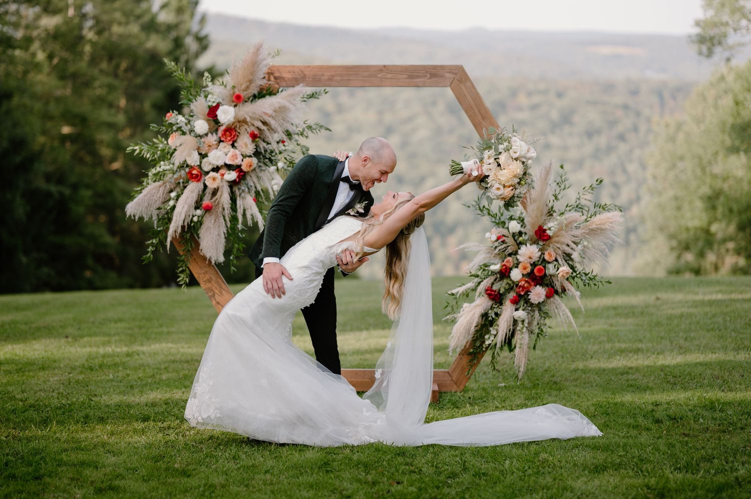 Groom dips bride in white gown before hexagonal floral arch with mountain backdrop.