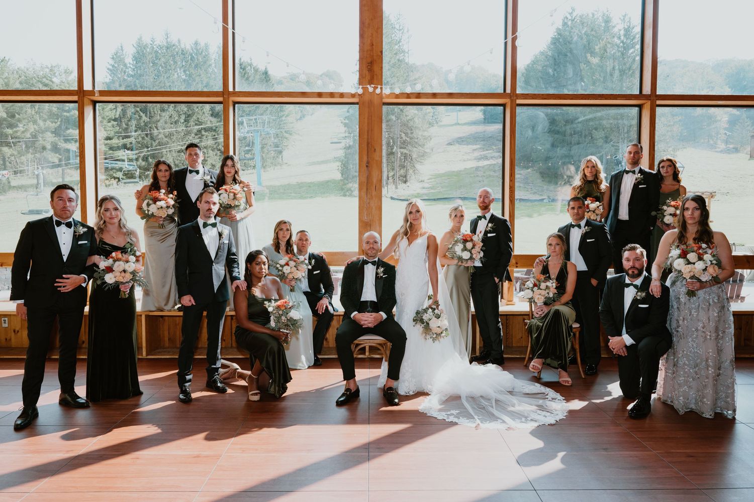 Wedding party group portrait in front of large windows with scenic outdoor view.