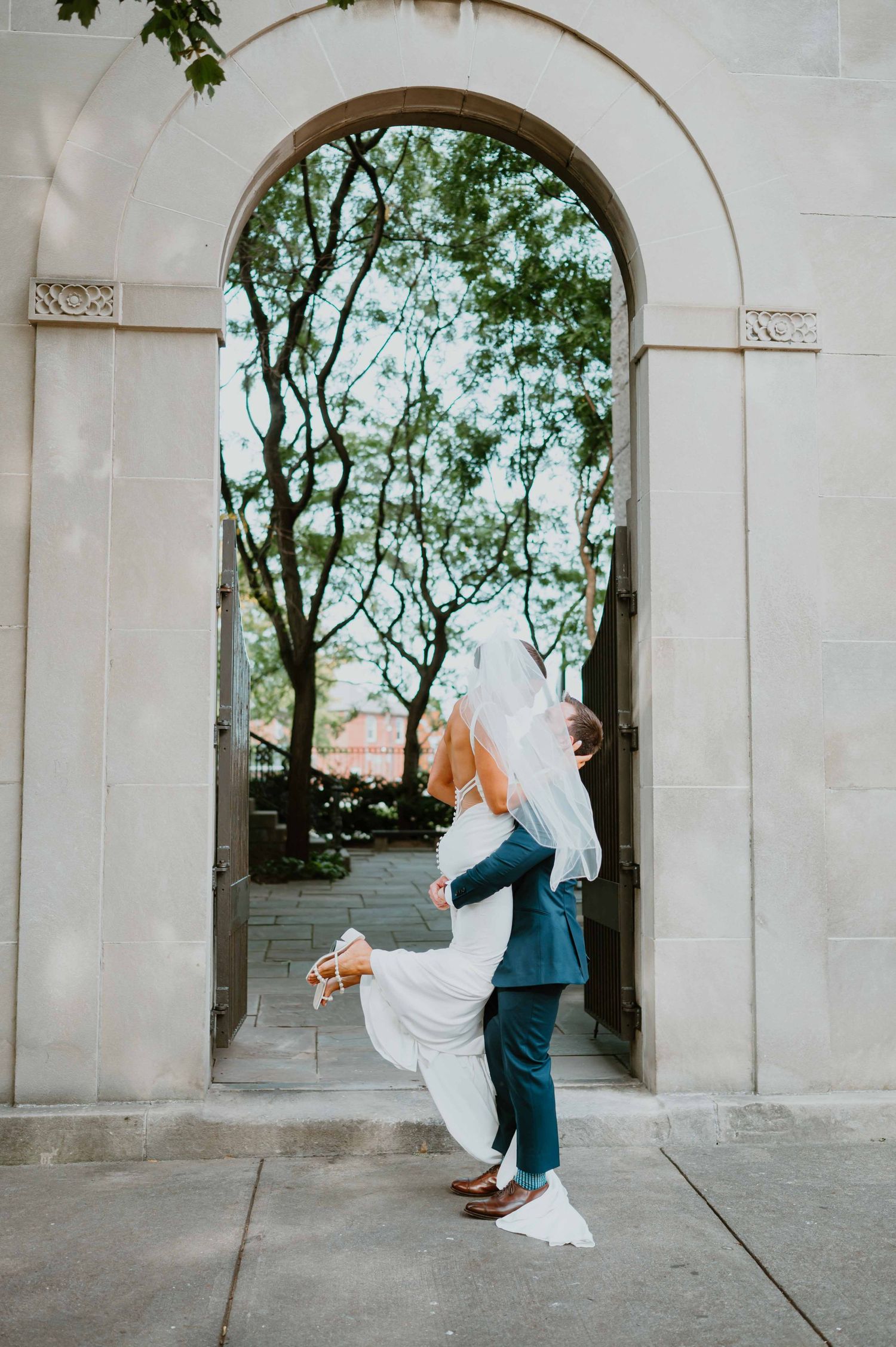 Groom lifts bride in a white gown for a kiss beneath an ornate stone archway surrounded by green trees.