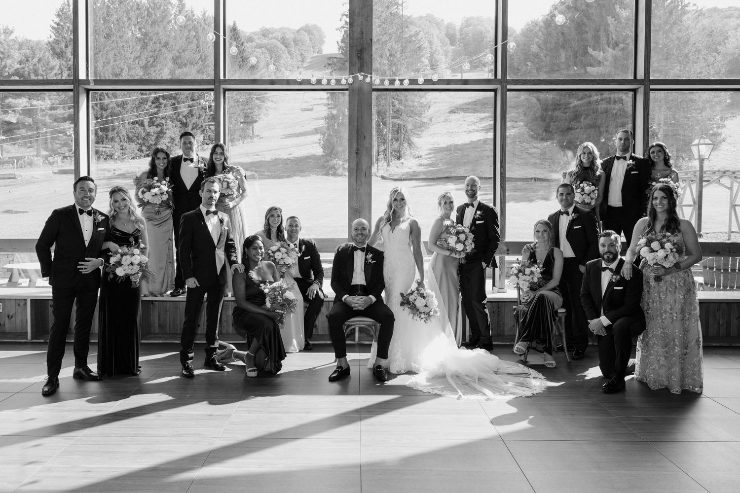 Large wedding party poses in black and white photo before panoramic windows.