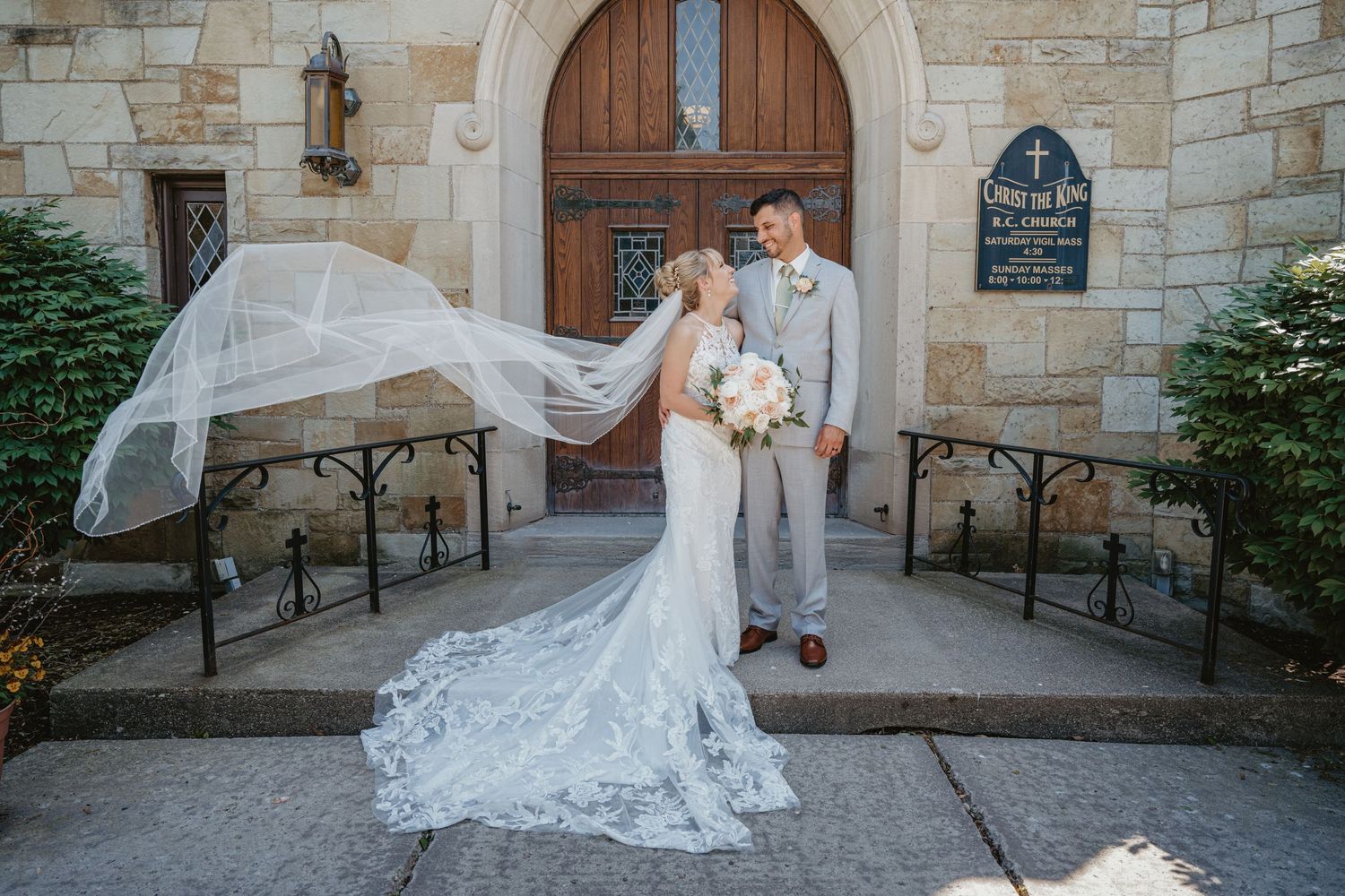 Bride's veil dramatically flows in the wind as couple poses before a stone church entrance on their wedding day.