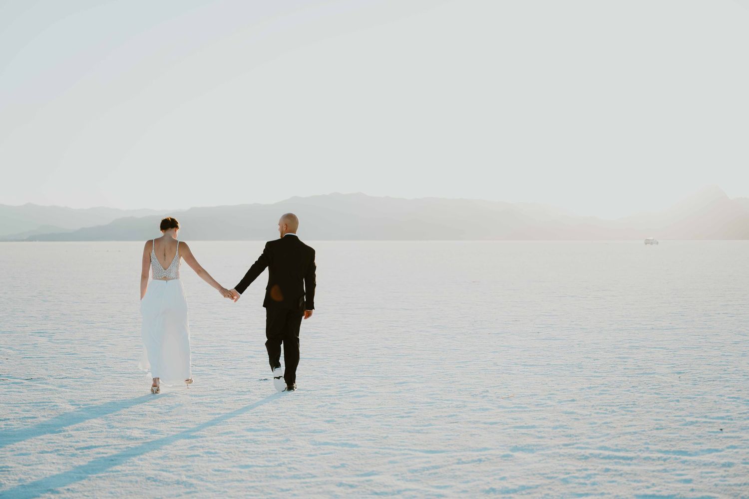 Bride and groom walk hand in hand across vast white salt flats at sunrise.
