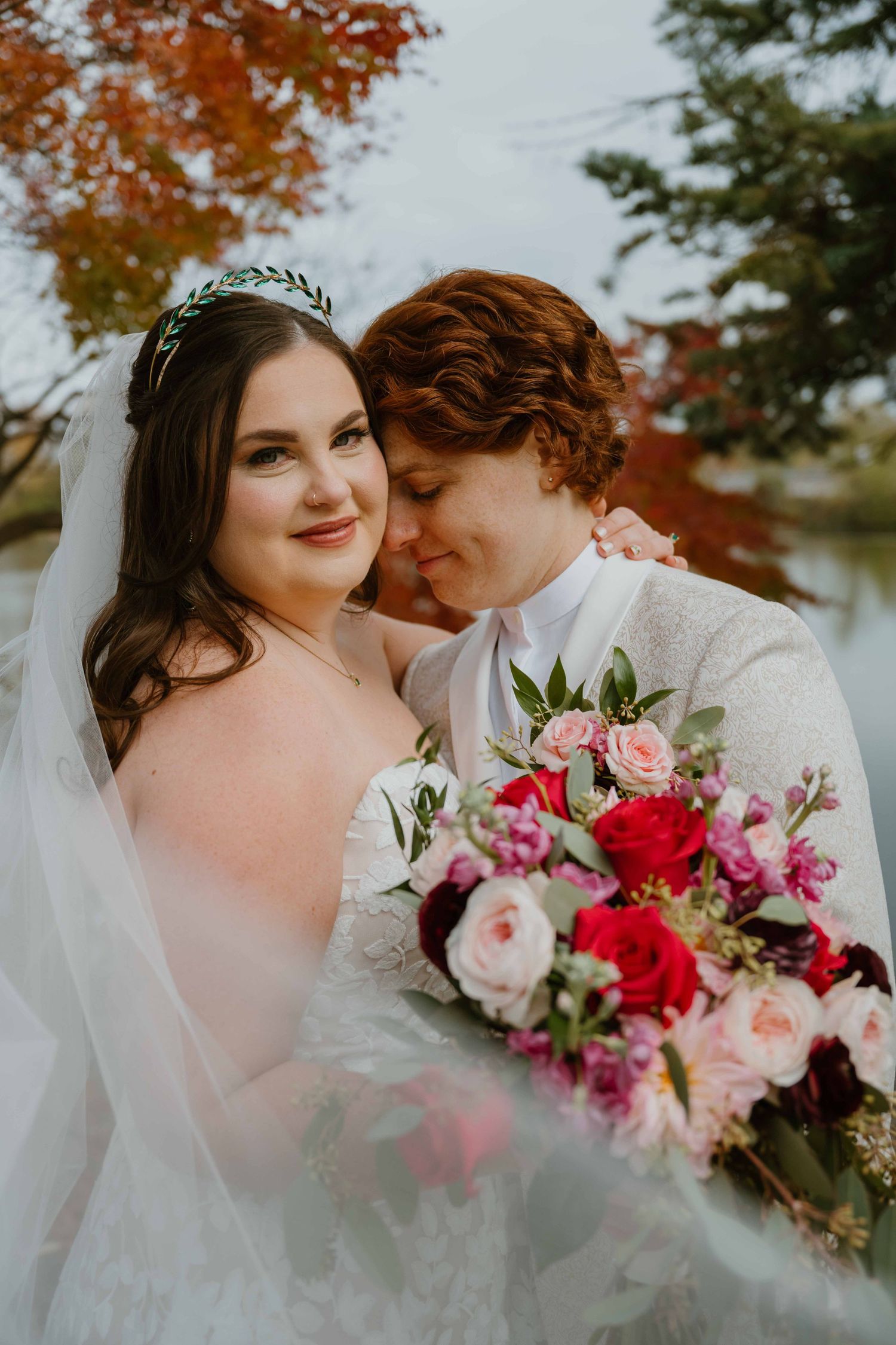 Two brides embrace outdoors in autumn, one holding a lush pink and red bouquet with fall foliage behind them.