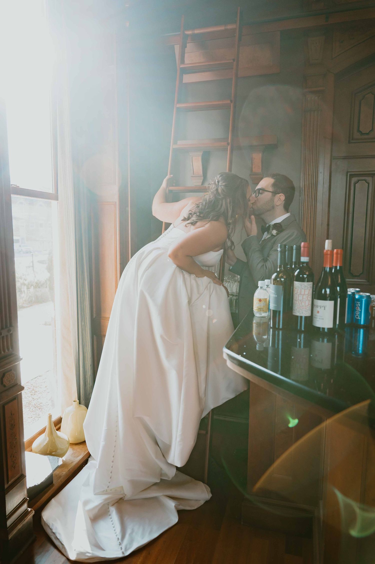 Bride and groom share a playful kiss near a wooden library ladder and wine bottles in soft light.