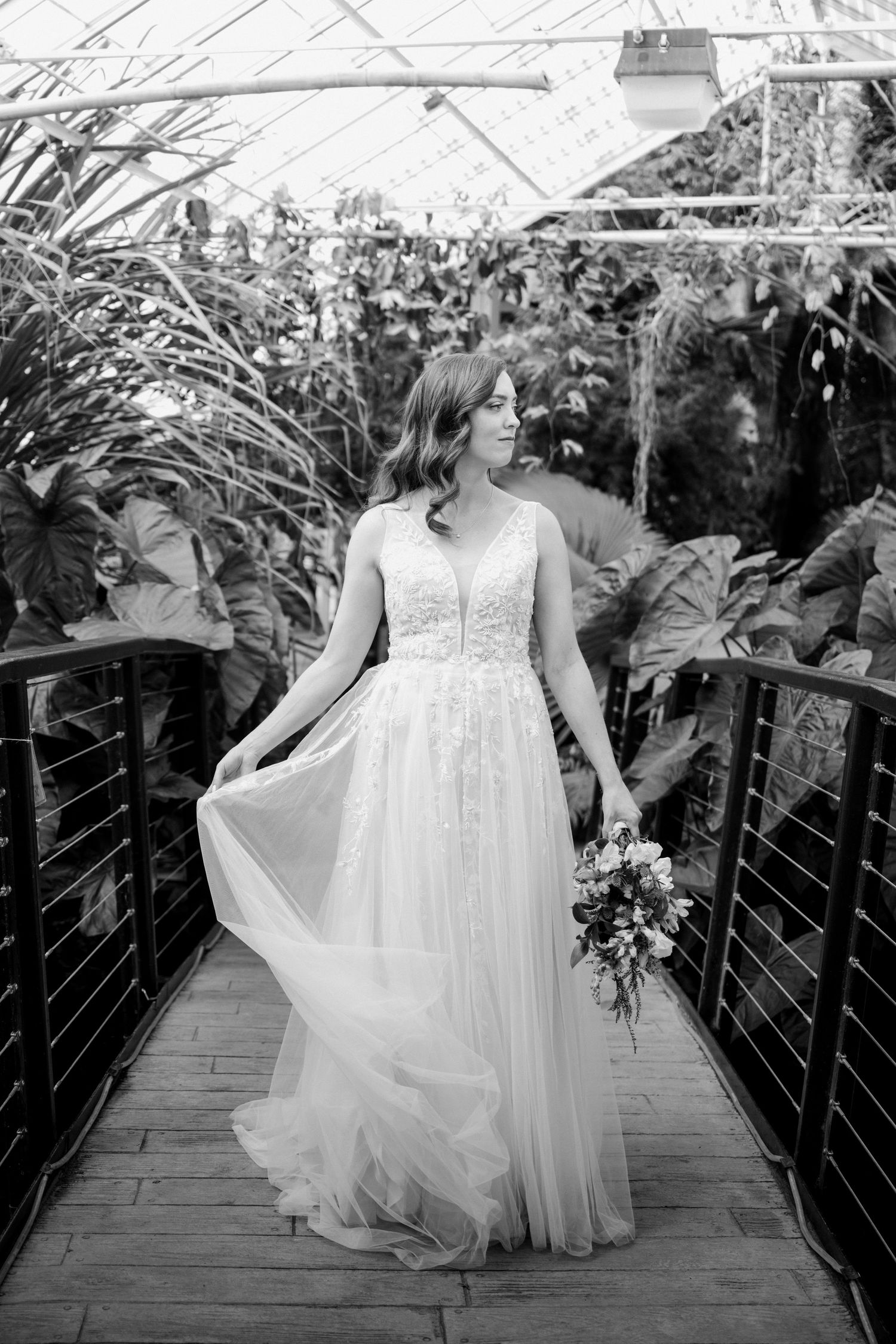 Black and white photo of bride in lace tulle gown walking on wooden bridge in botanical greenhouse.
