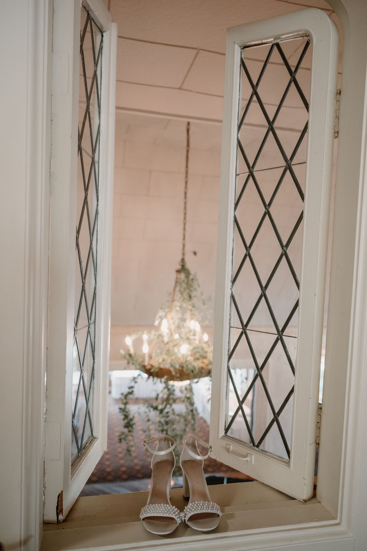 Elegant bridal heels displayed on a windowsill with a chandelier reflected in diamond-paned glass doors.