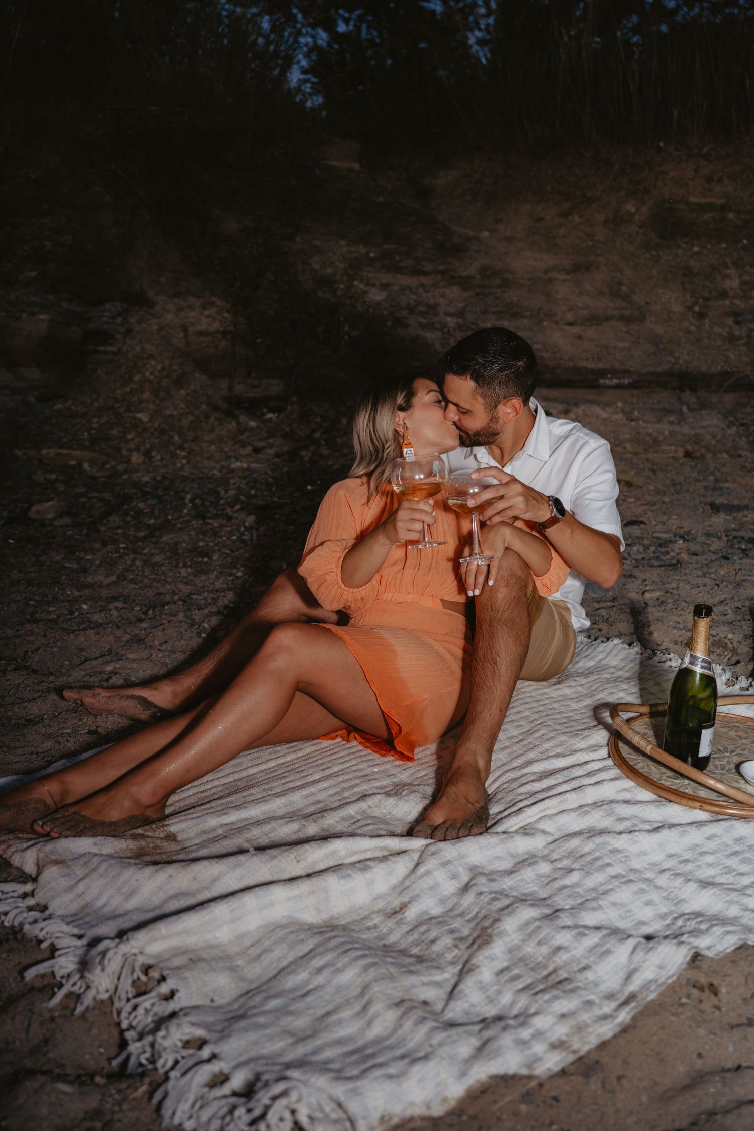 Couple kissing during a romantic beach picnic at dusk, holding champagne glasses on a blanket.