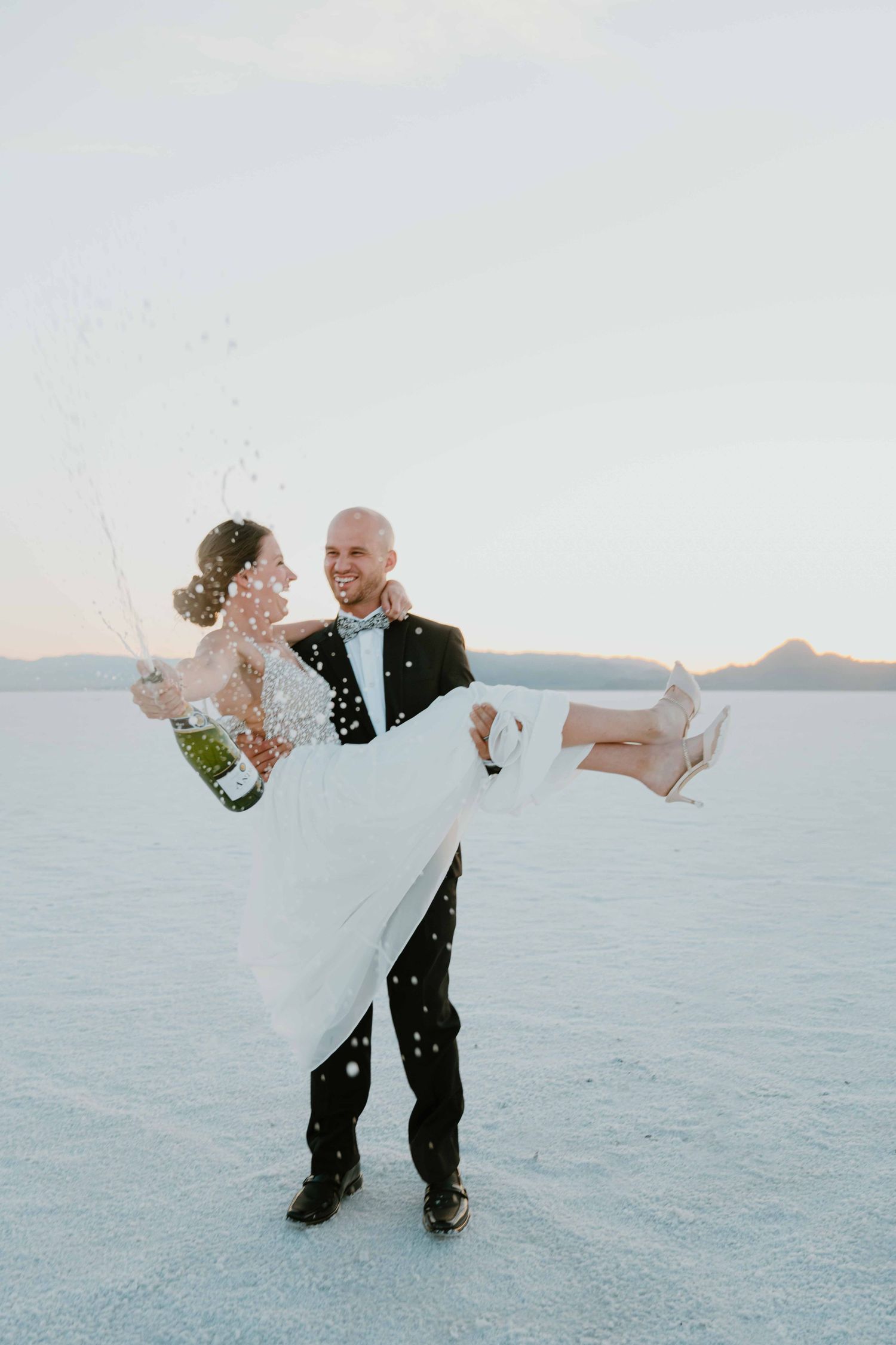 Groom lifts smiling bride while champagne sprays in the air on the bright white salt flats.