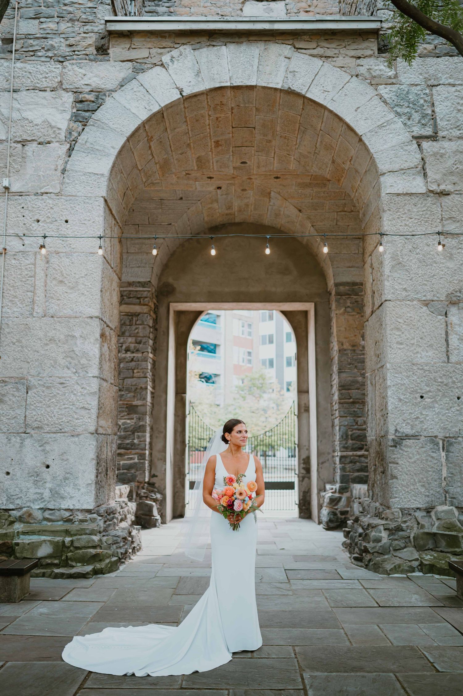 Bride in elegant white gown with train stands under stone archway at historic venue.