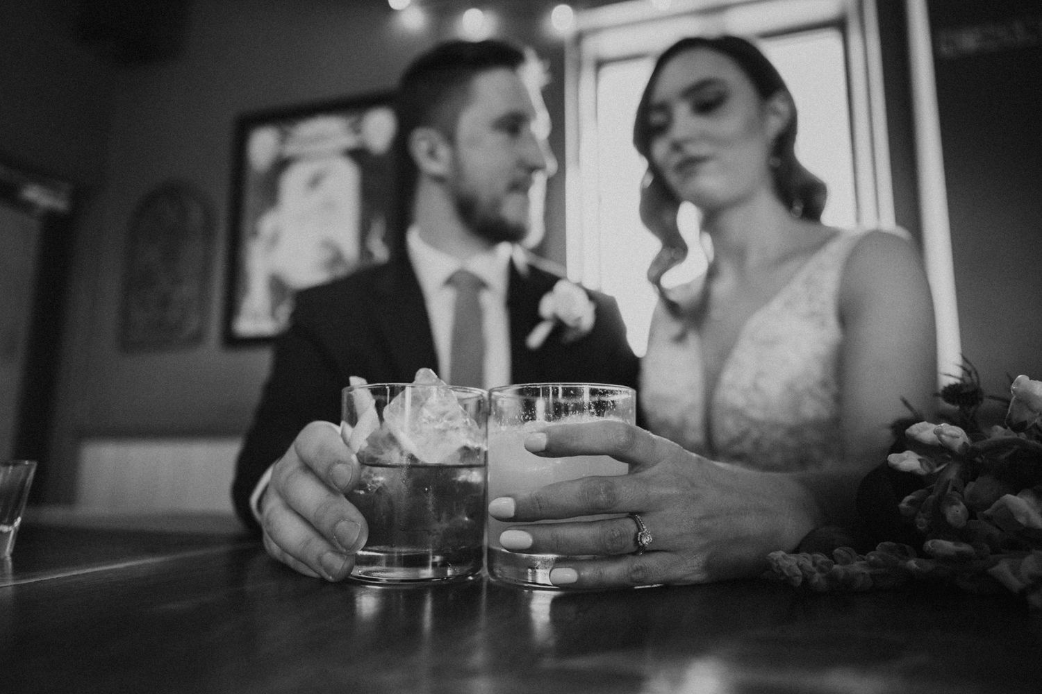 Black and white close-up of bride and groom clinking cocktail glasses at a bar on wedding day.