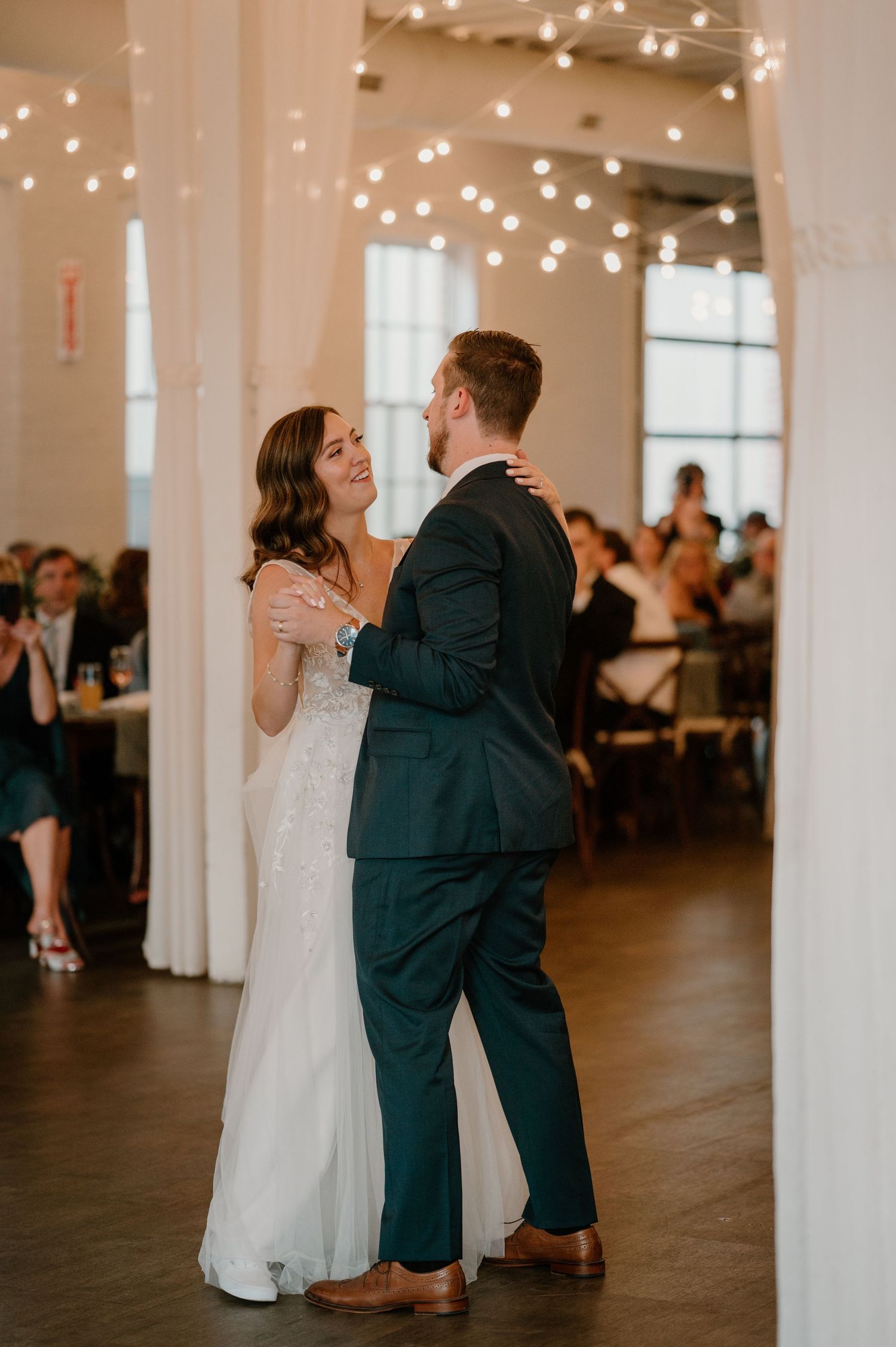 Bride smiles joyfully at groom during first dance under string lights at wedding reception.