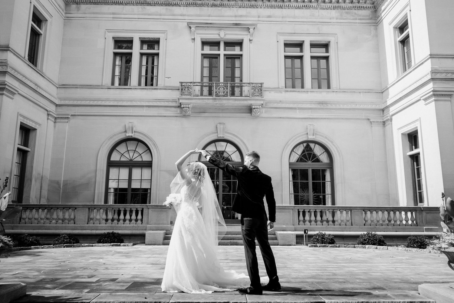 Black and white photo of bride and groom embracing before grand historic mansion.