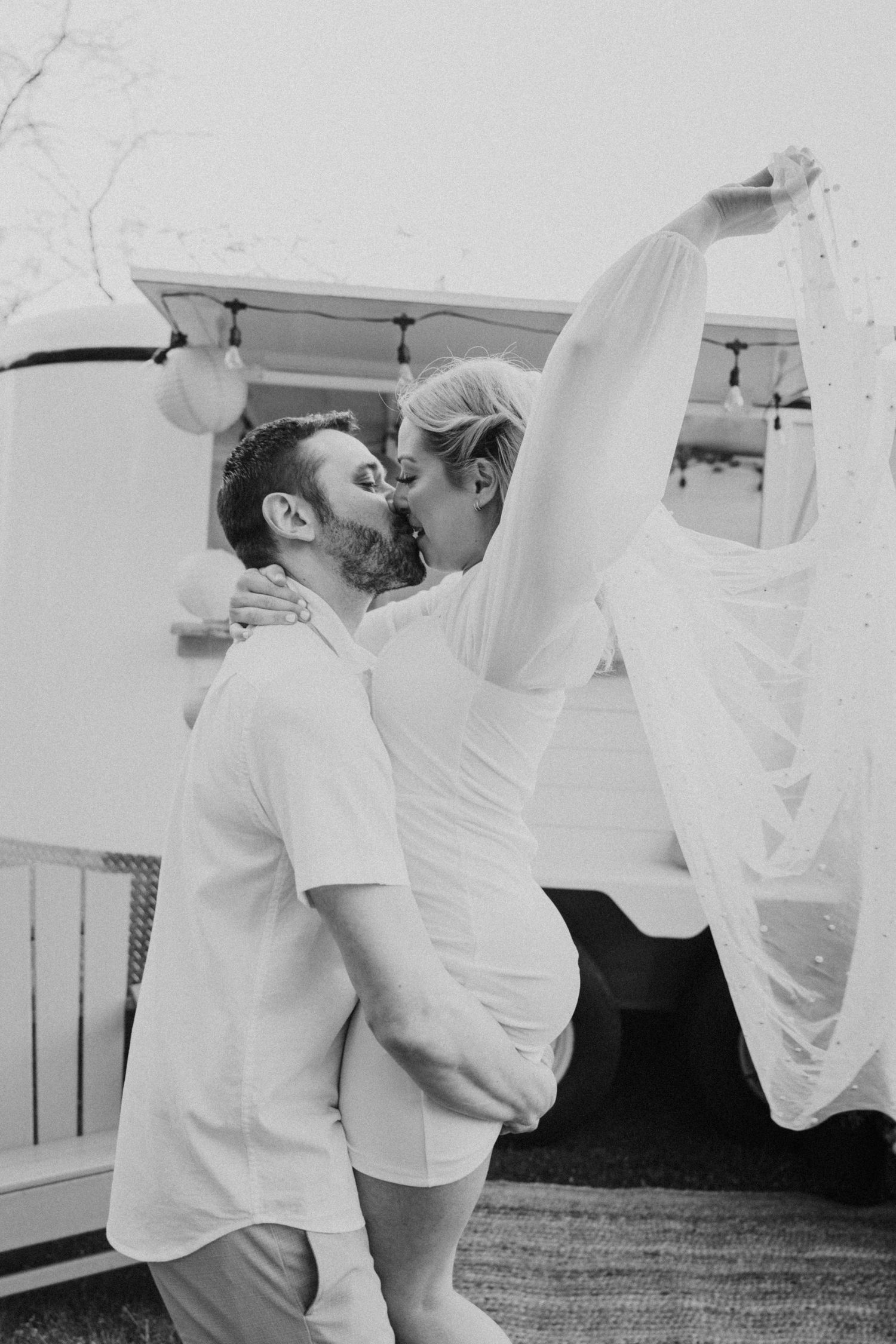 Black and white photo of couple kissing while groom lifts bride in wedding dress near white truck.