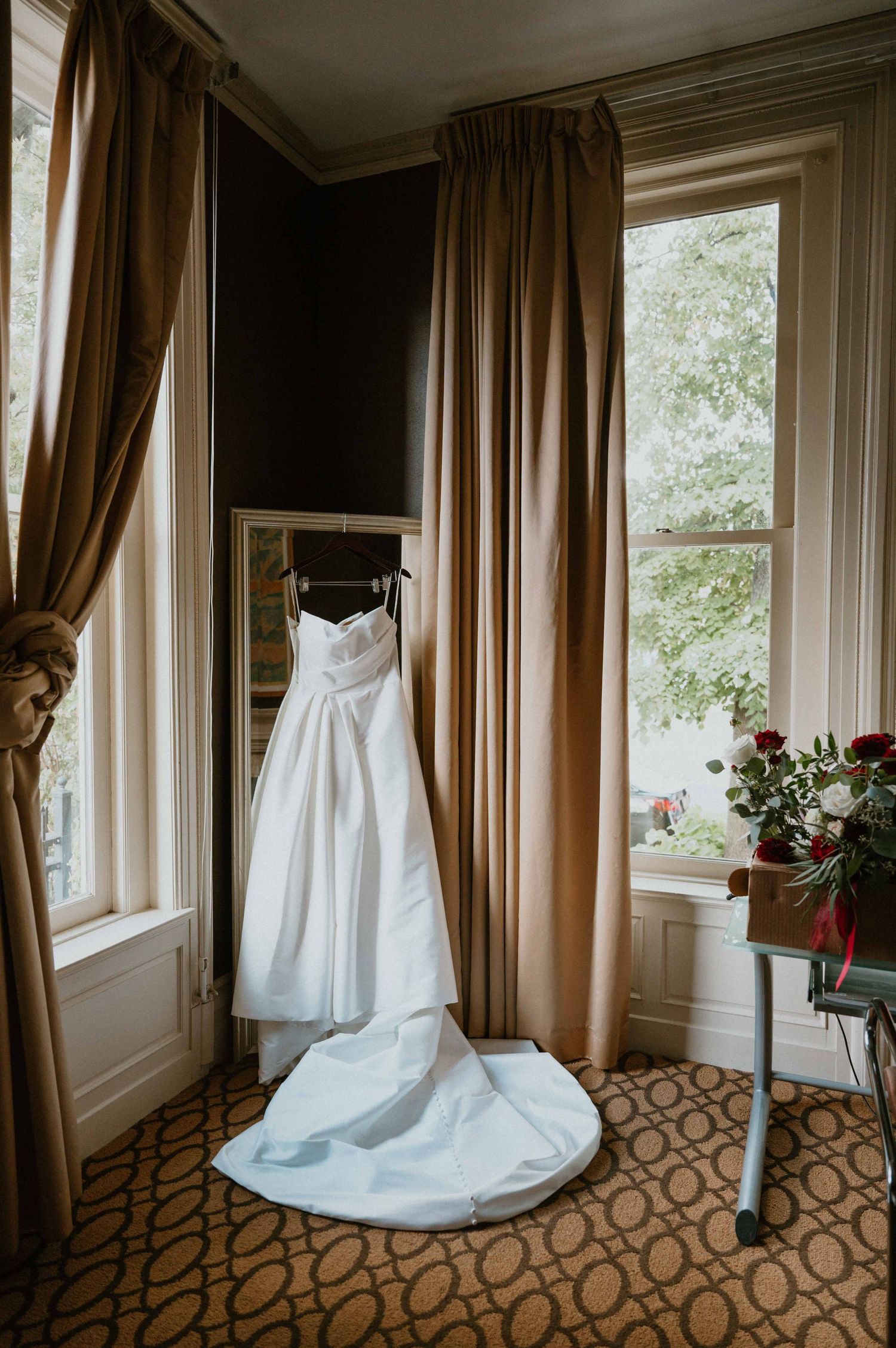 Elegant strapless white wedding gown with train displayed hanging between golden curtains by a window.