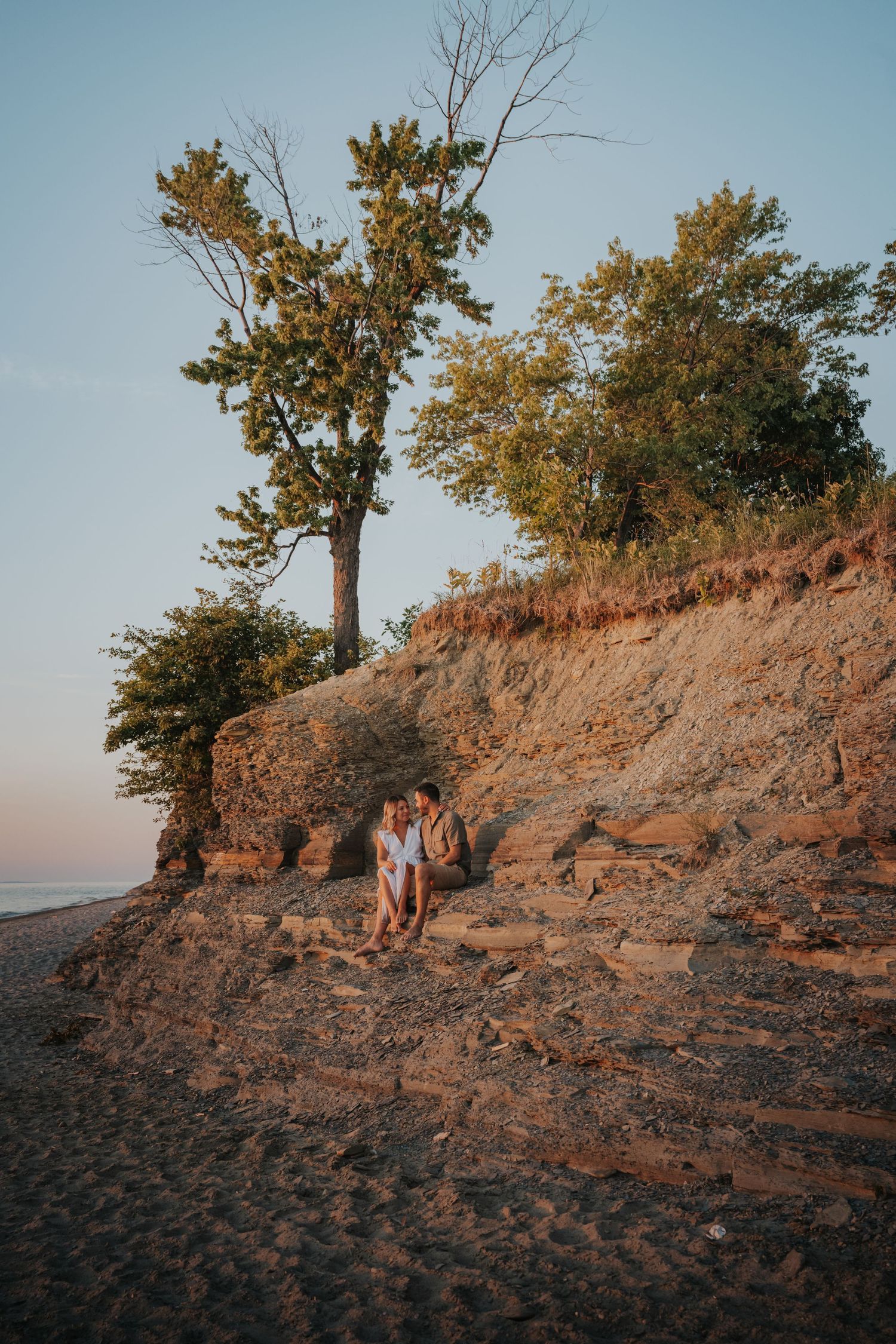 Couple sits closely together on a sandy beach cliff at sunset surrounded by lush green trees.