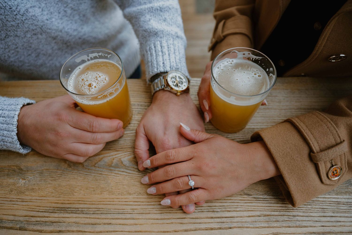 Close-up of engaged couple holding craft beers on wooden table, showing engagement ring and watch.