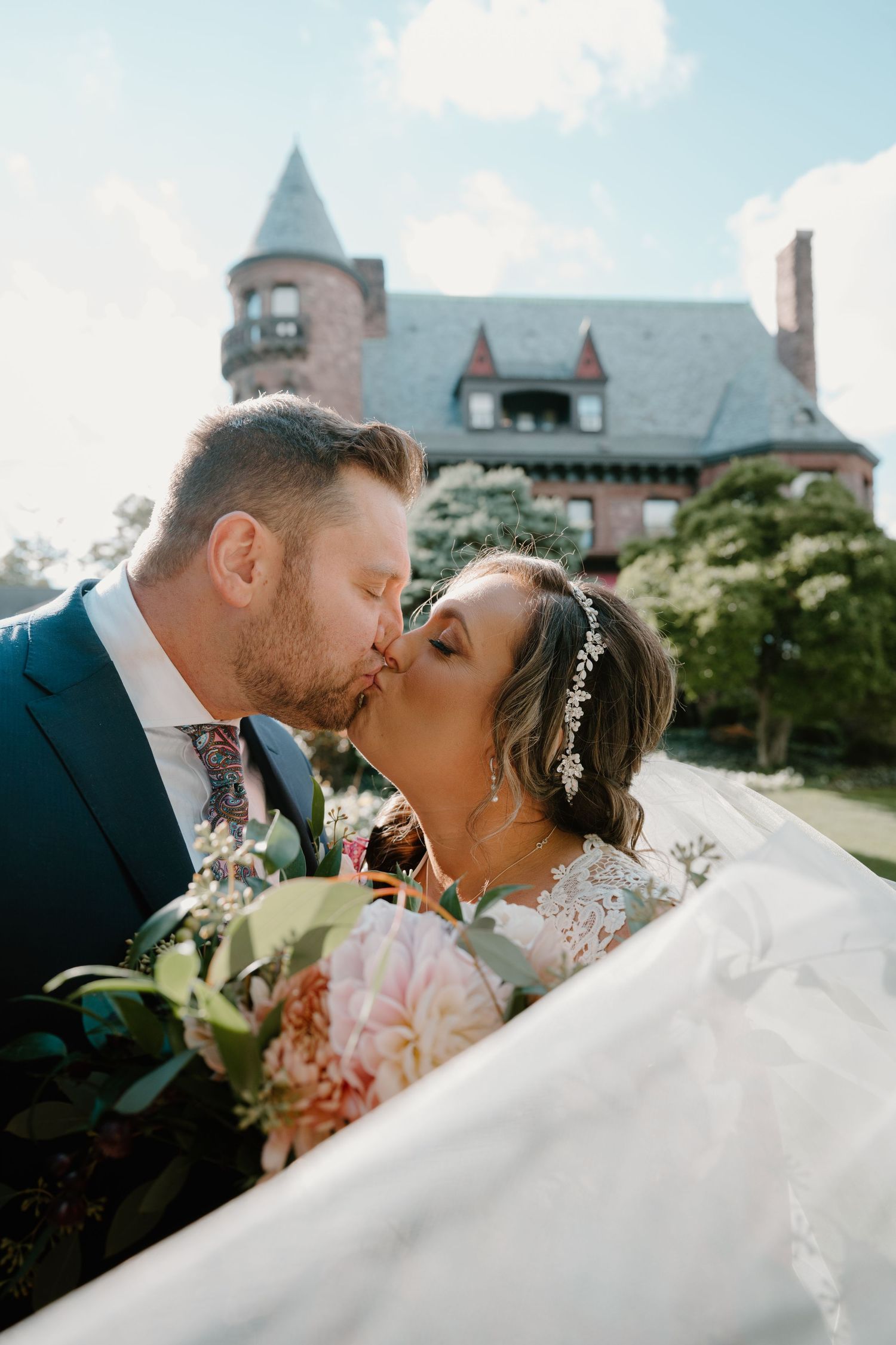 Bride and groom sharing a romantic kiss outdoors with a historic castle-like building in the background.