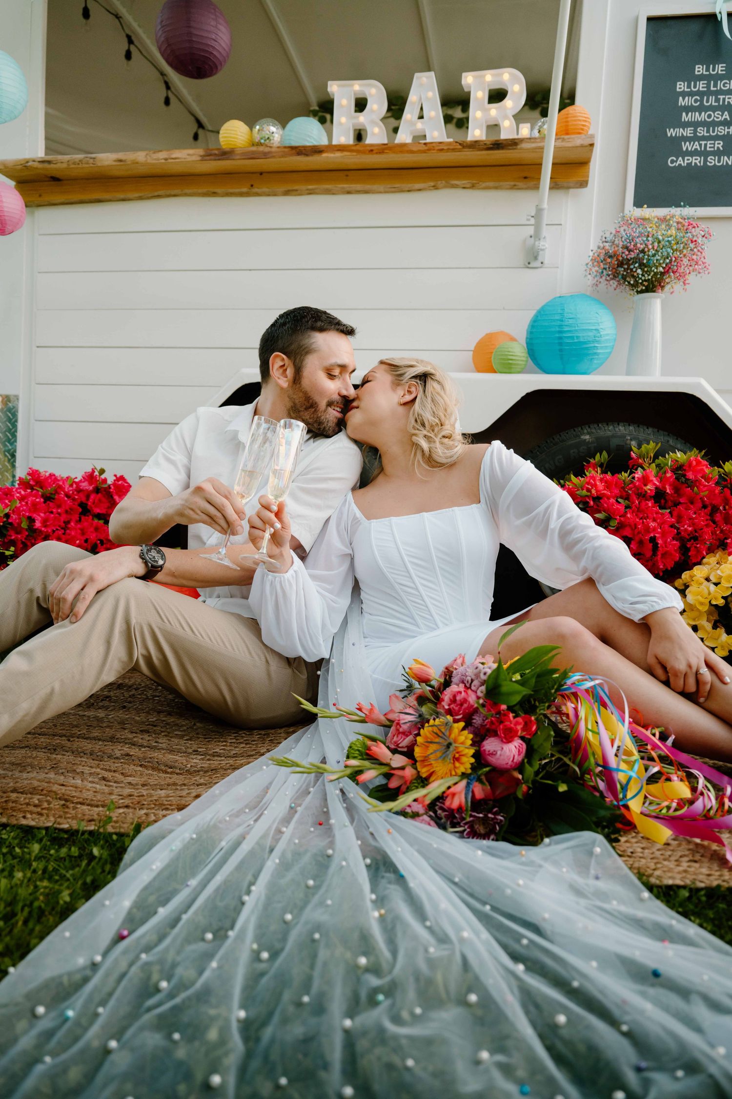 Bride and groom toast champagne while sitting on ground before a BAR truck with colorful flowers.